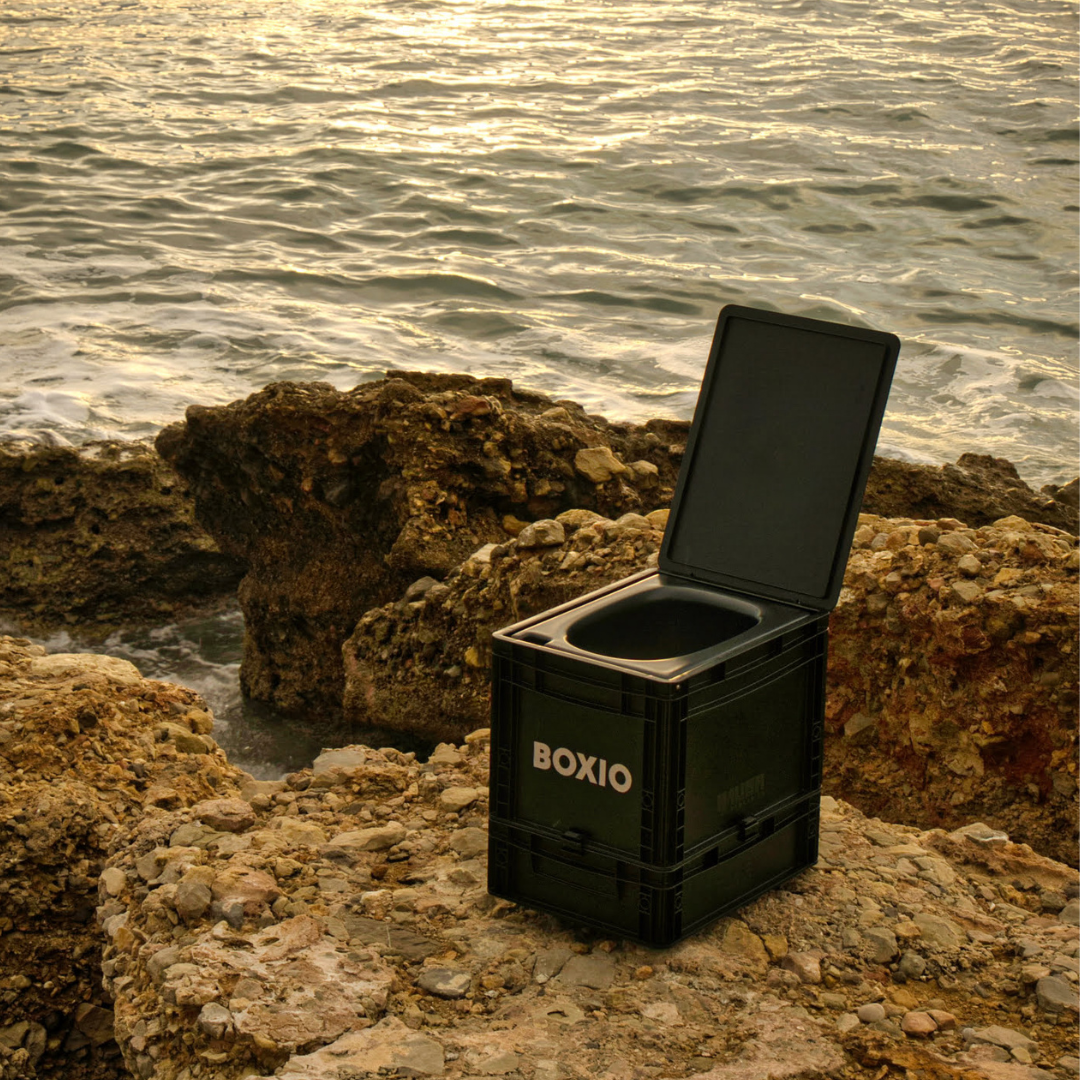 Schwarze BOXIO-Box mit offenem Deckel, auf Steinen am Ufer, im Hintergrund glitzerndes Wasser.