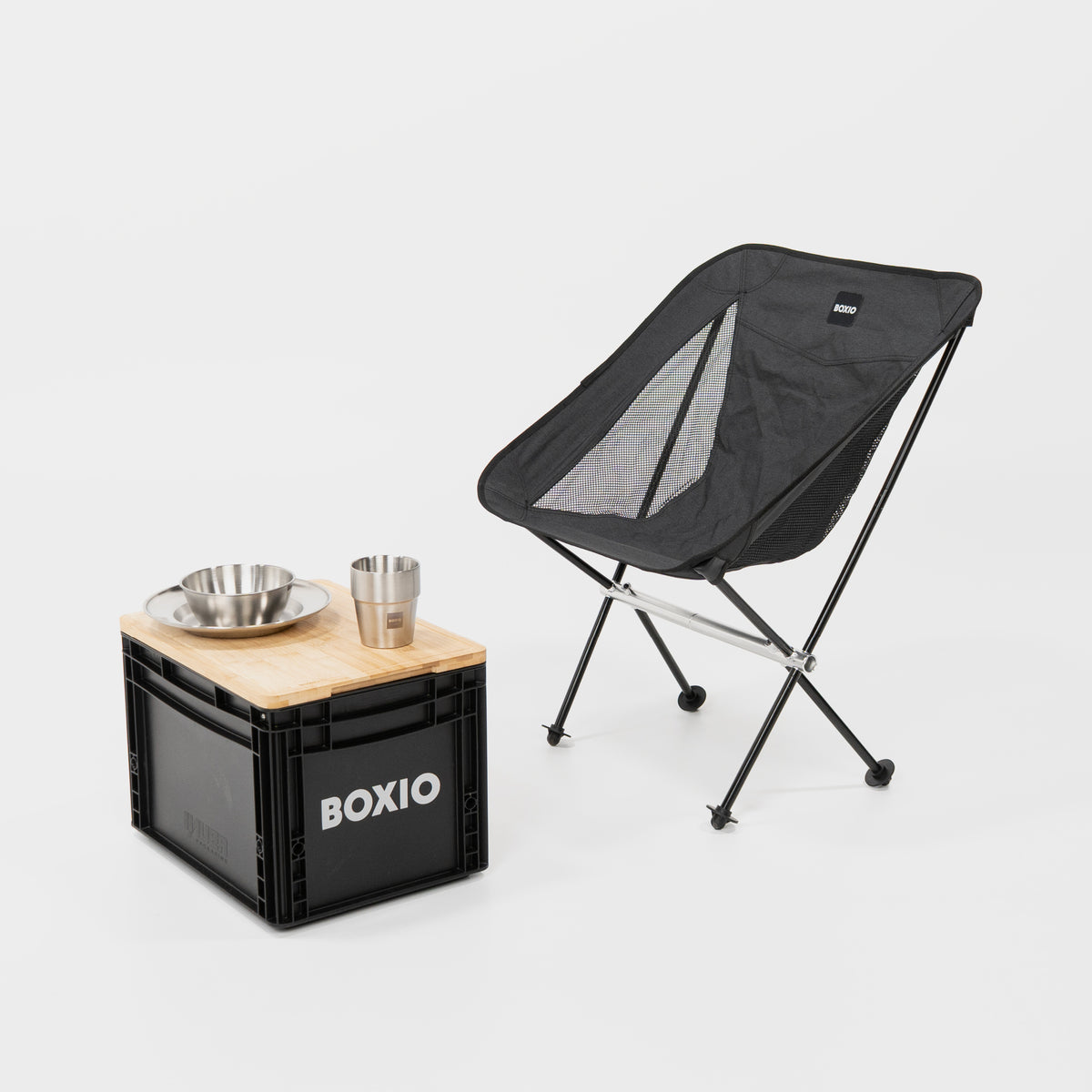 Campingstuhl mit seitlichen Lüftungsschlitzen im Bezug. Davor eine BOXIO Eurobox mit Schneidebrett auf dem Deckel. Darauf das BOXIO Campinggeschirr vor weißem Hintergrund