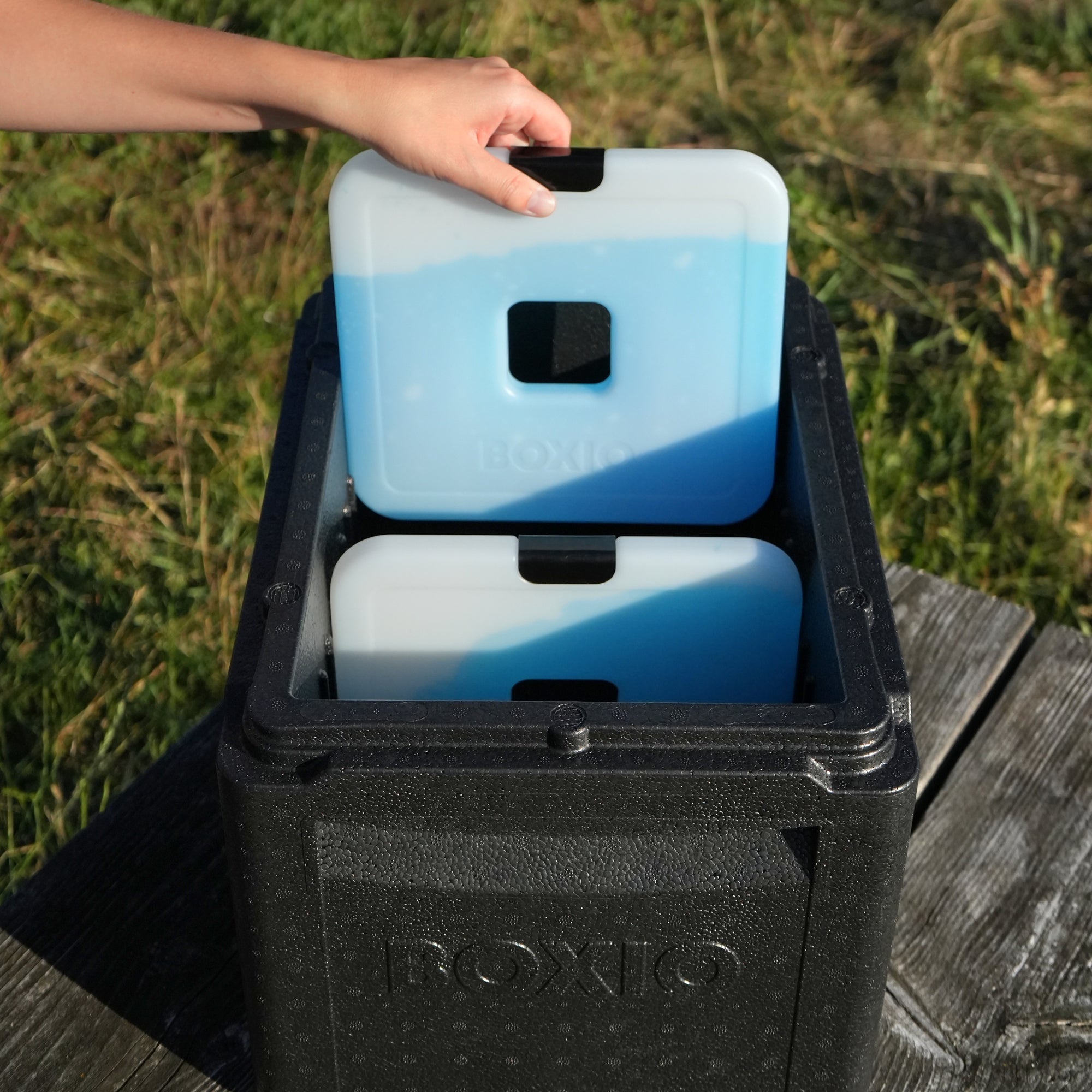 Hand setzt einen blauen Kühlakku in eine schwarze Kühlbox, die auf einer Holzfläche steht.