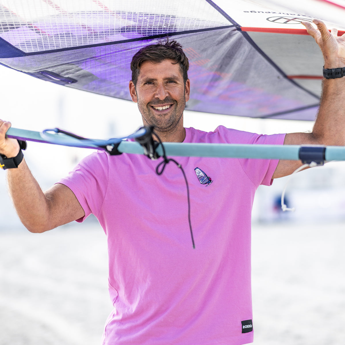 Lächelnder Windsurfer trägt ein Segel und zeigt seine Ausrüstung am Strand, im Hintergrund sind unscharfe Sandflächen.