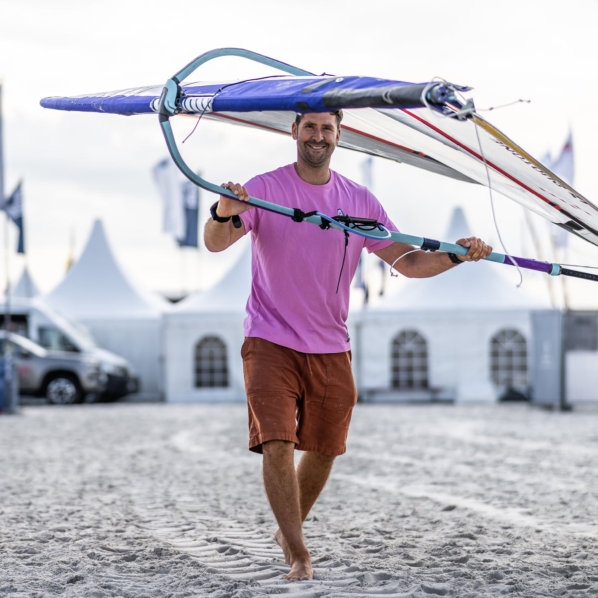 Mann trägt ein Windsurfbrett am Strand, trägt ein pinkes T-Shirt und kurze Hosen, lächelt und geht auf sandigem Untergrund.