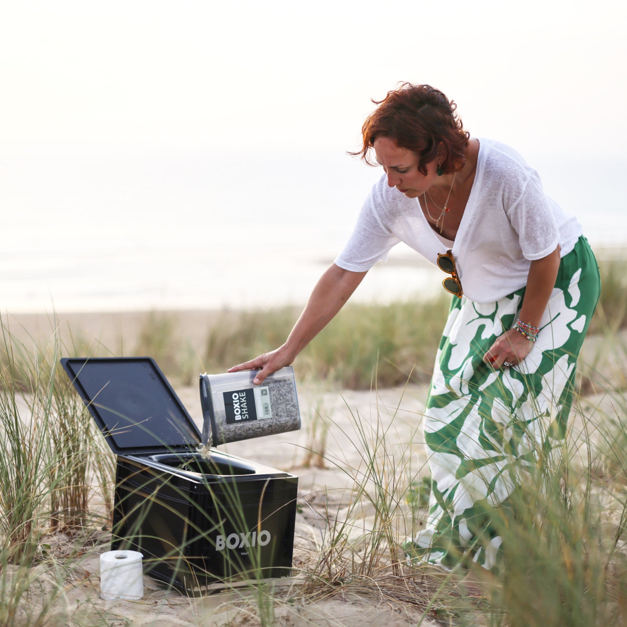 Frau in Strandkleidung gießt einen Behälter in eine schwarze Box im Sand, umgeben von Gräsern und Küstenlandschaft.