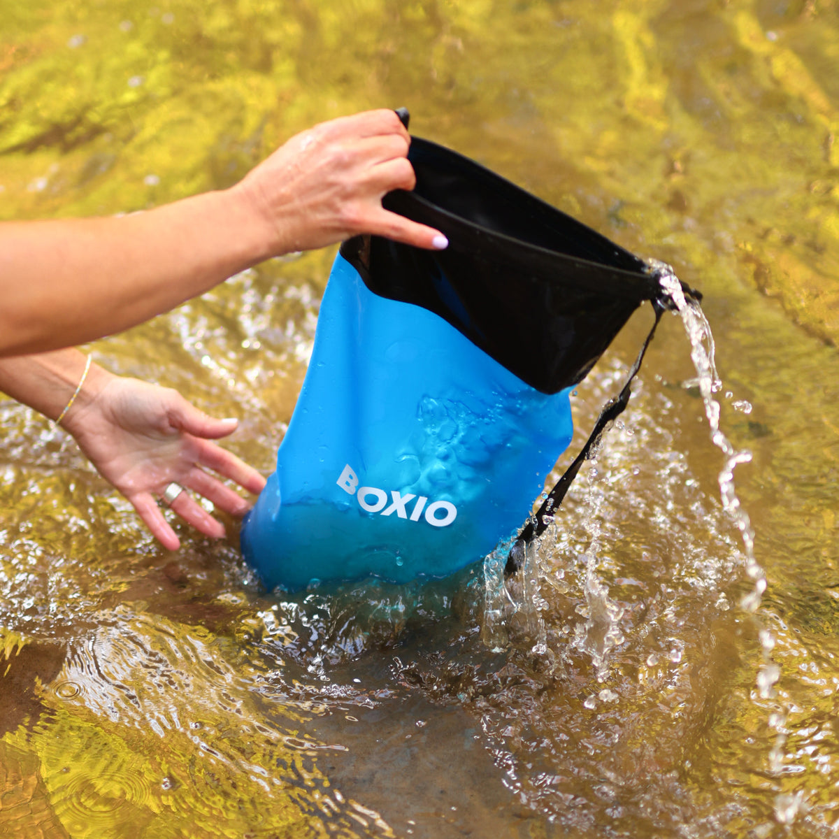 Hand greift in eine wasserdichte, blaue Boxio-Tasche, die Wasser ausgießt, im klaren Flusswasser.