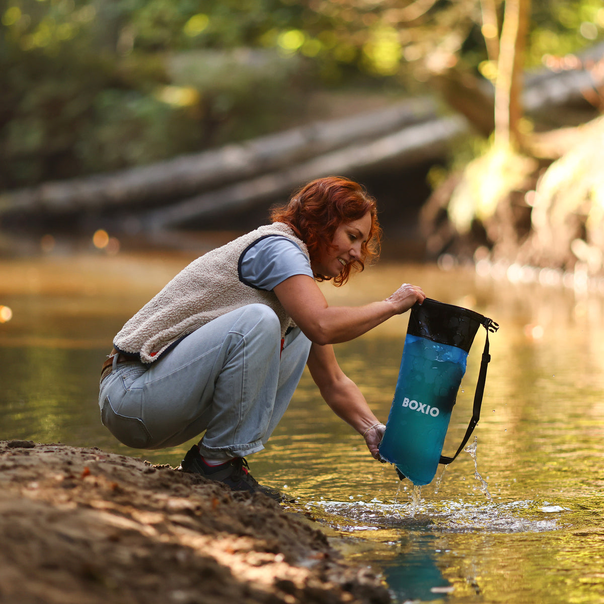 Frau mit rotem Haar beugt sich über einen Fluss und füllt einen blauen Wasserfilterbehälter.