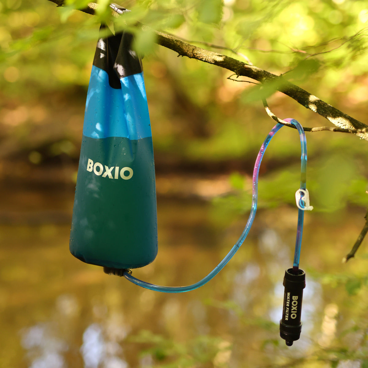 Eine blaue Wasserfilter-Tasche hängt an einem Ast, verbunden mit einem Schlauch und einem Wasserfilter.