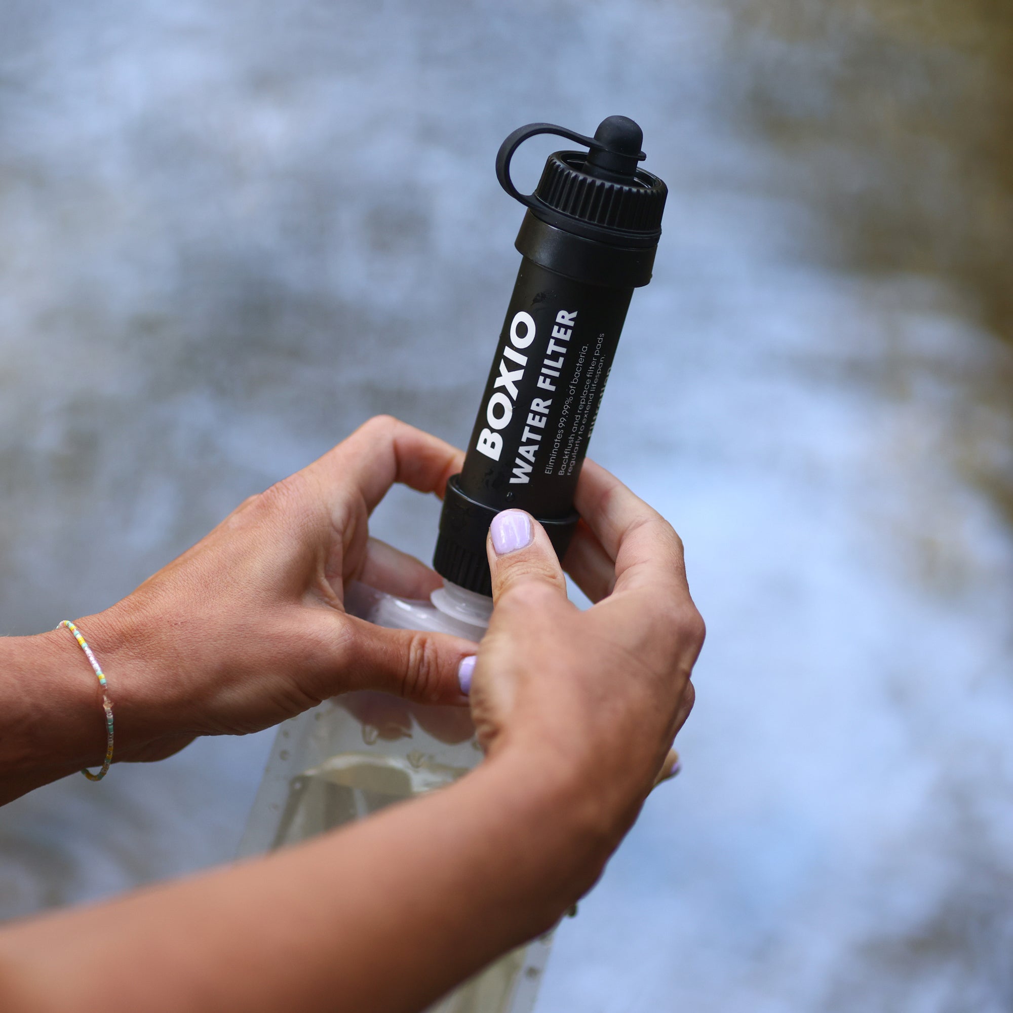 Hände setzen einen Boxio-Wasserfilter auf eine transparente Wasserflasche am Ufer eines Gewässers.