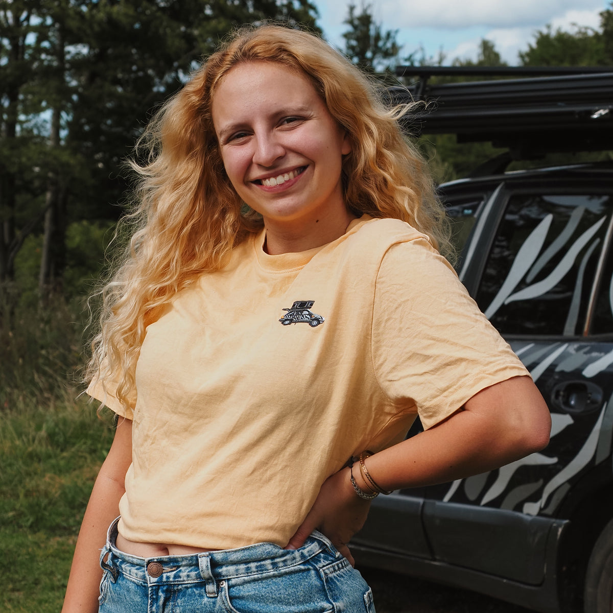 Junge Frau mit lockigem, blondem Haar, lächelnd in einem gelben T-Shirt und Jeans, im Freien vor einem Auto.