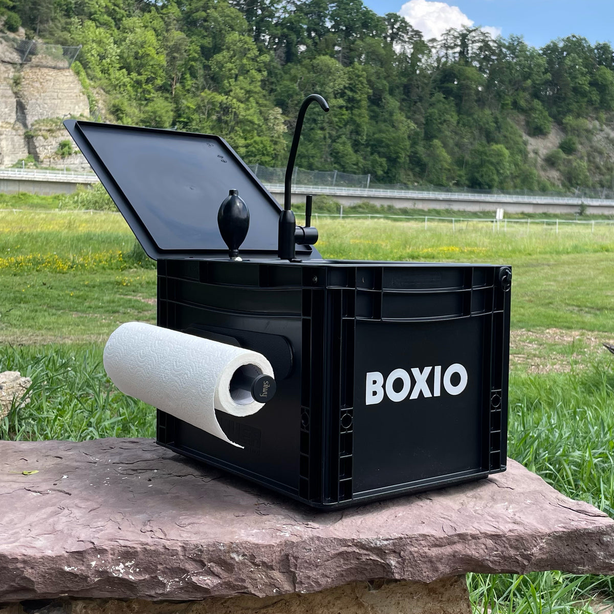 Schwarze Boxio-Box mit herausragender Küchenrolle und geöffnetem Deckel auf einem Felsen im Freien.