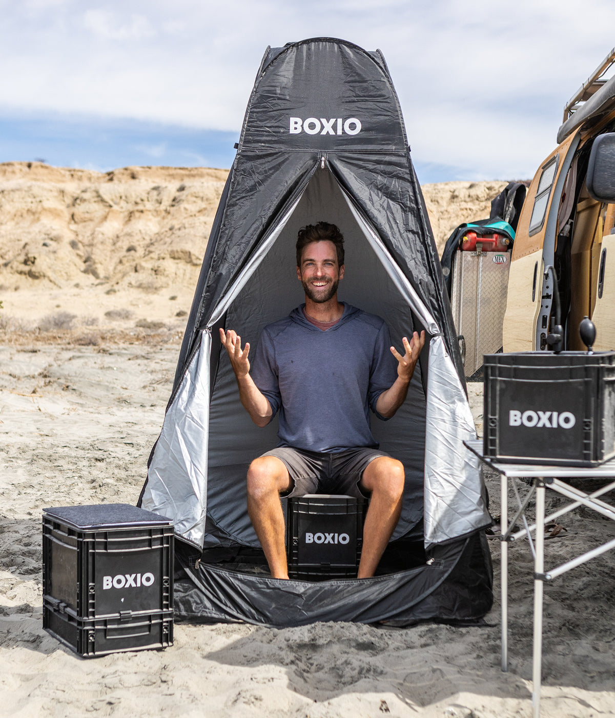 Mann sitzt in einem schwarzen Campingzelt mit "BOXIO"-Logo, umgeben von Campingboxen und einem Geländewagen.