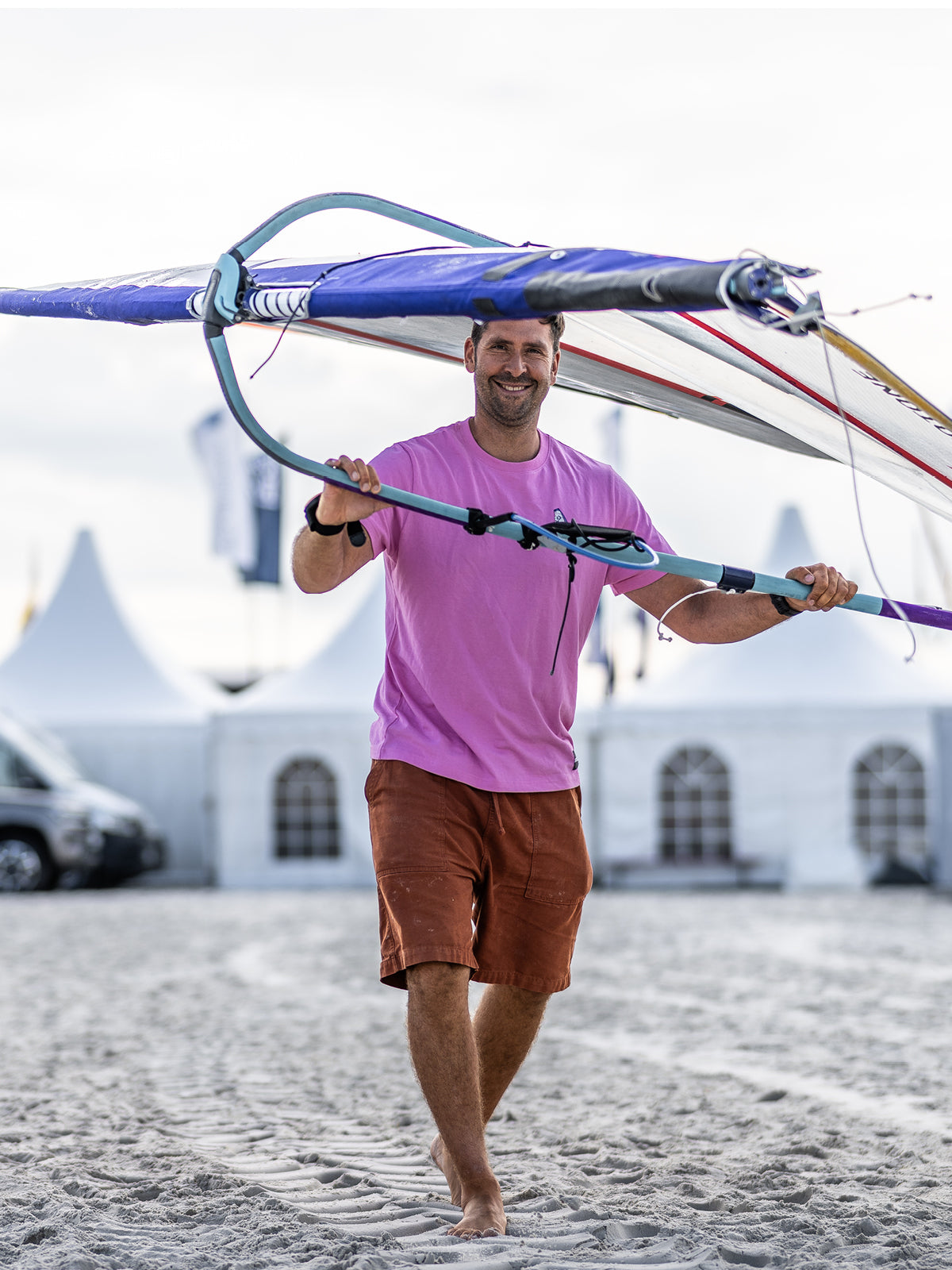 Mann in pinkem Shirt trägt ein Windsurfbrett über den Strand, Zelte im Hintergrund.