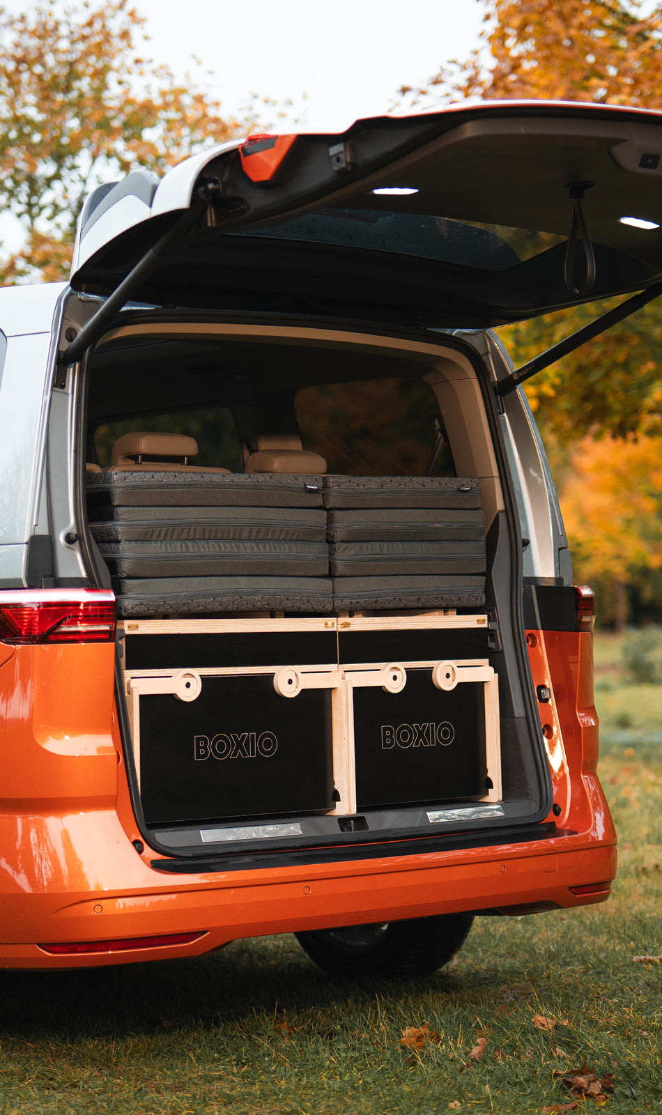 Heckansicht eines orangefarbenen Vans mit geöffnetem Kofferraum, ausgestattet mit einer Holzbox und Matratzen.