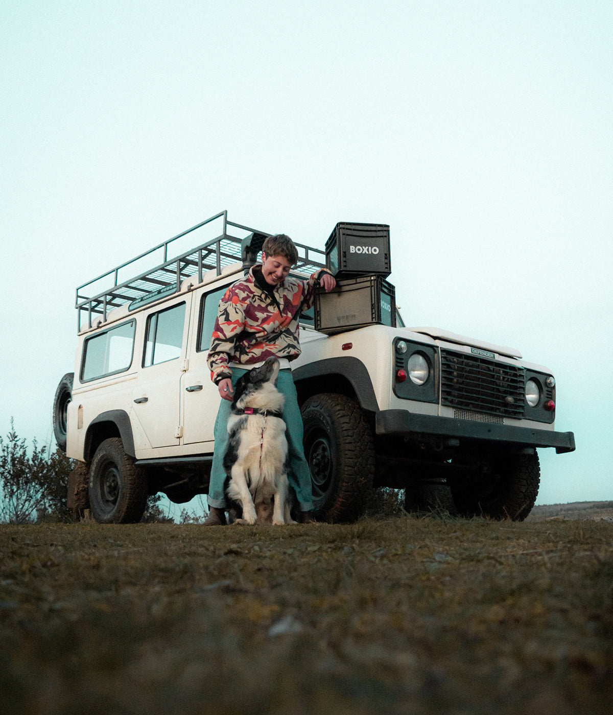 Junger Mann mit Camouflagejacke steht neben einem Geländewagen und spielt mit einem Hund auf einer Wiese.