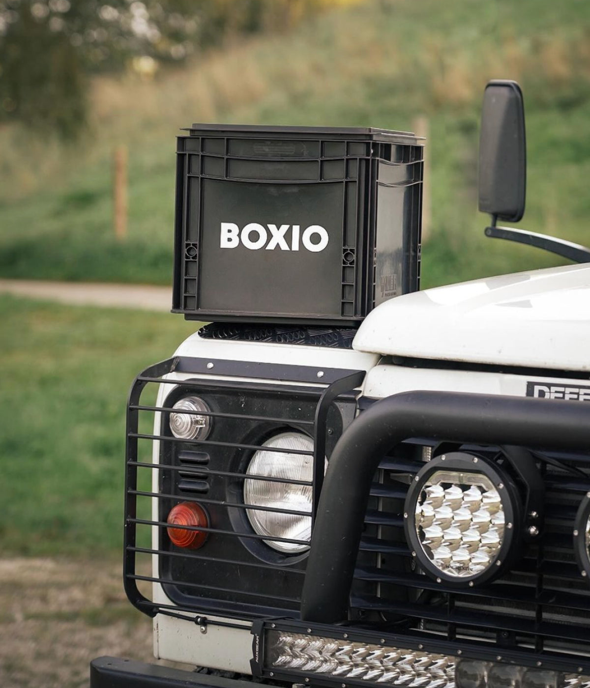 Schwarze BOXIO-Transportbox auf einem weißen Geländewagen mit Grill und Scheinwerfern im Vordergrund.