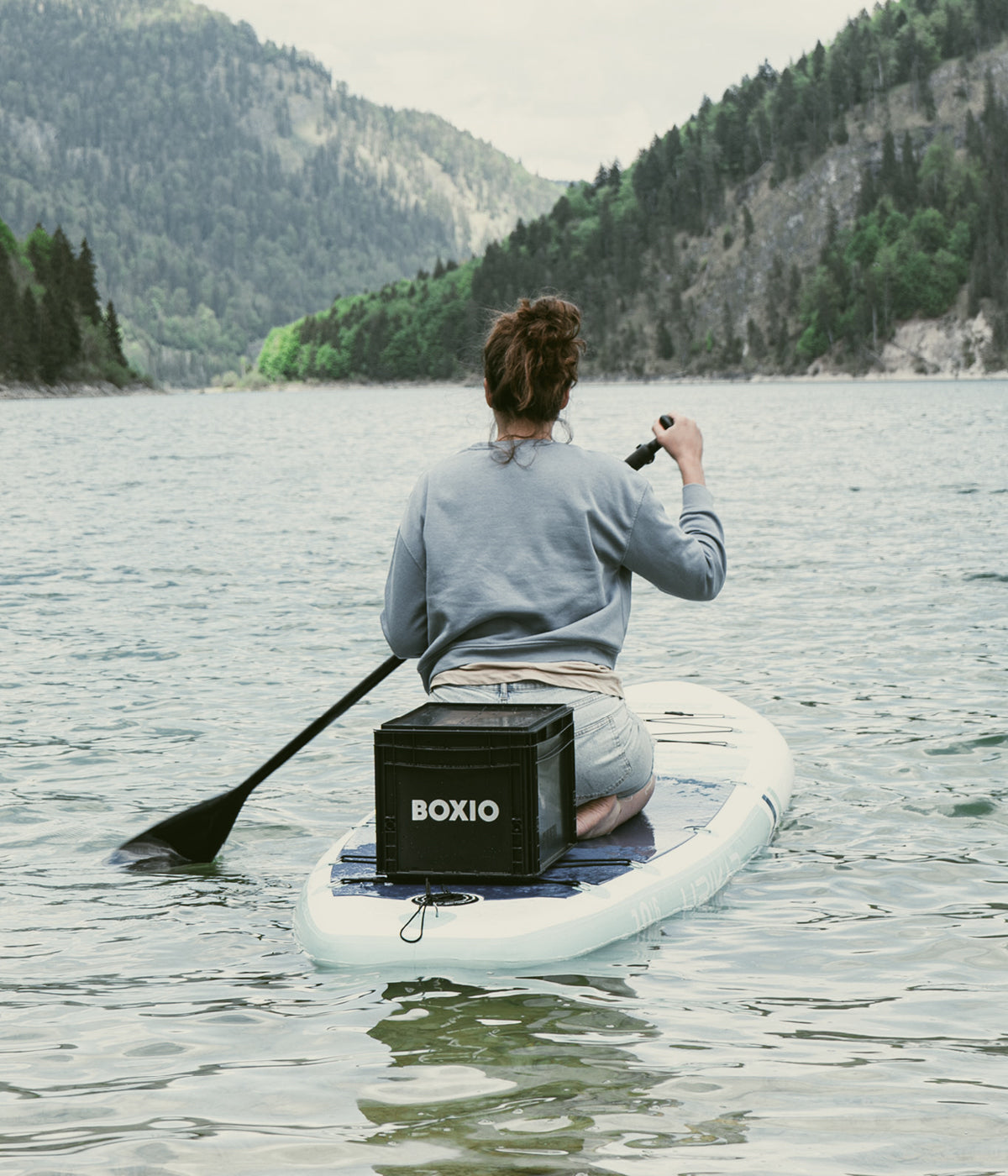 Frau paddelt auf einem Stand-Up-Paddle-Board in einem ruhigen Gewässer mit hügeliger, bewaldeter Landschaft im Hintergrund.