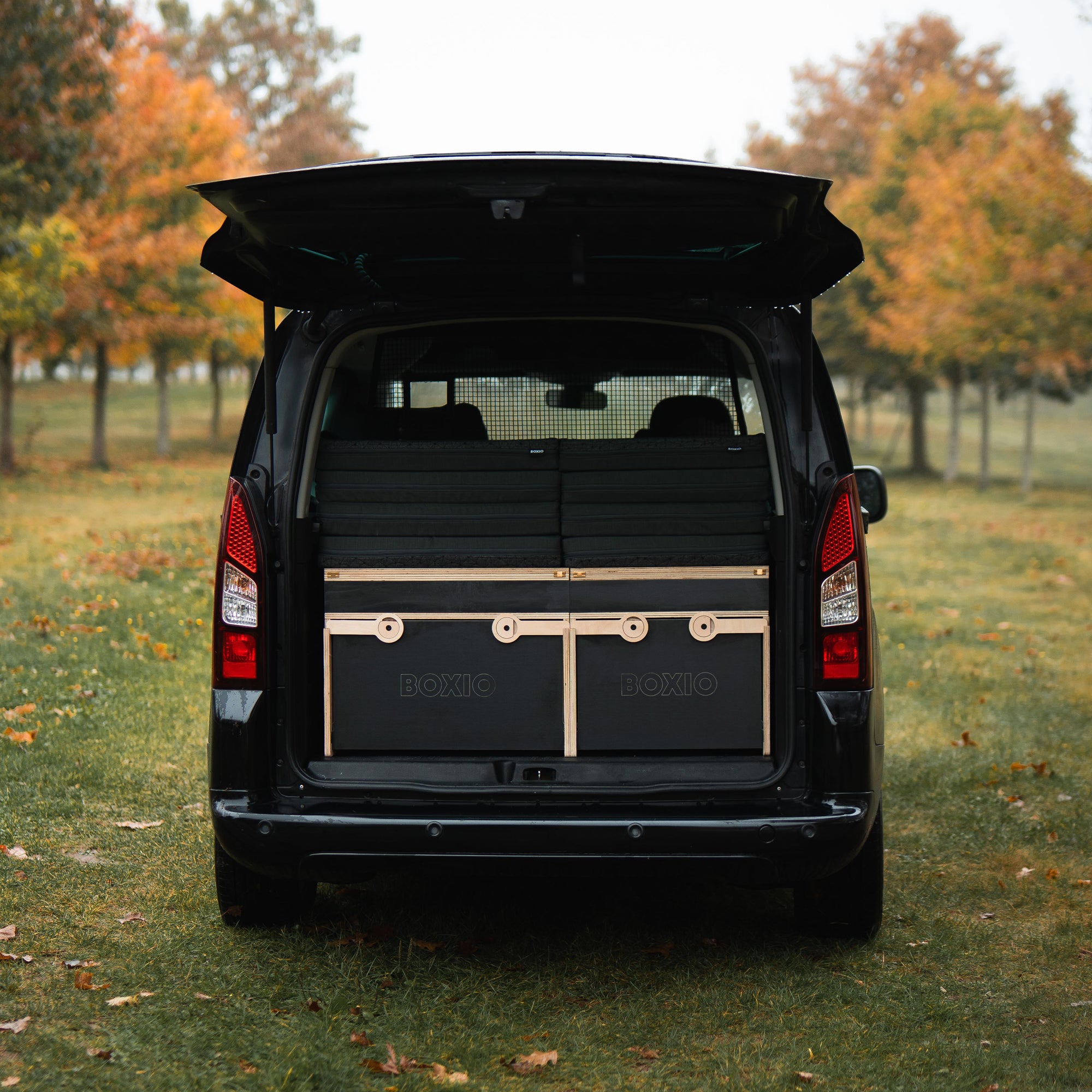 Heckansicht eines schwarzen Vans mit geöffnetem Kofferraum, ausgestattet mit Holzboxen und Sitzpolstern, umgeben von Bäumen.