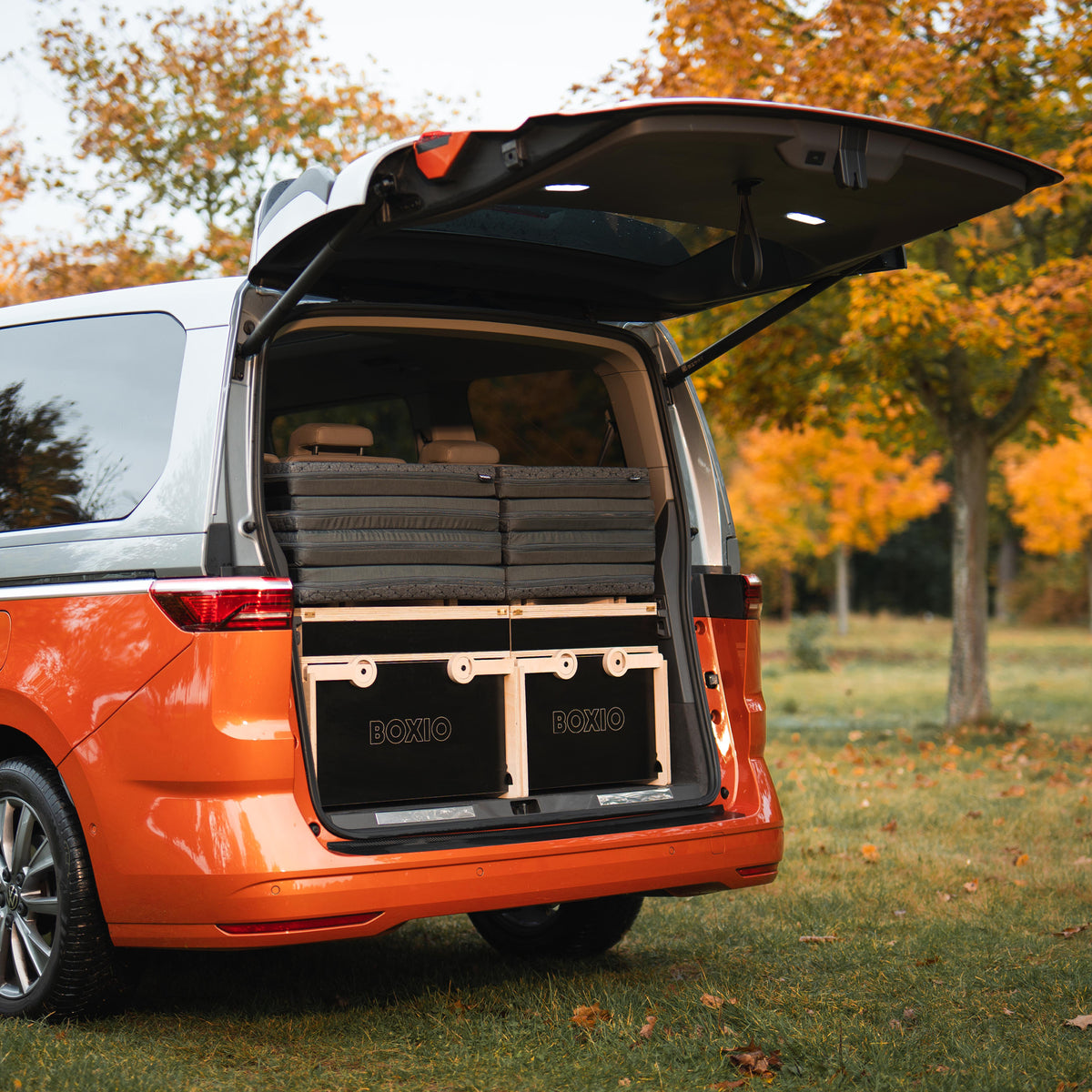 Offenes Kofferraum eines orangefarbenen Vans mit gestapelten Kisten und Holzplatten, Hintergrund mit Herbstbäumen.