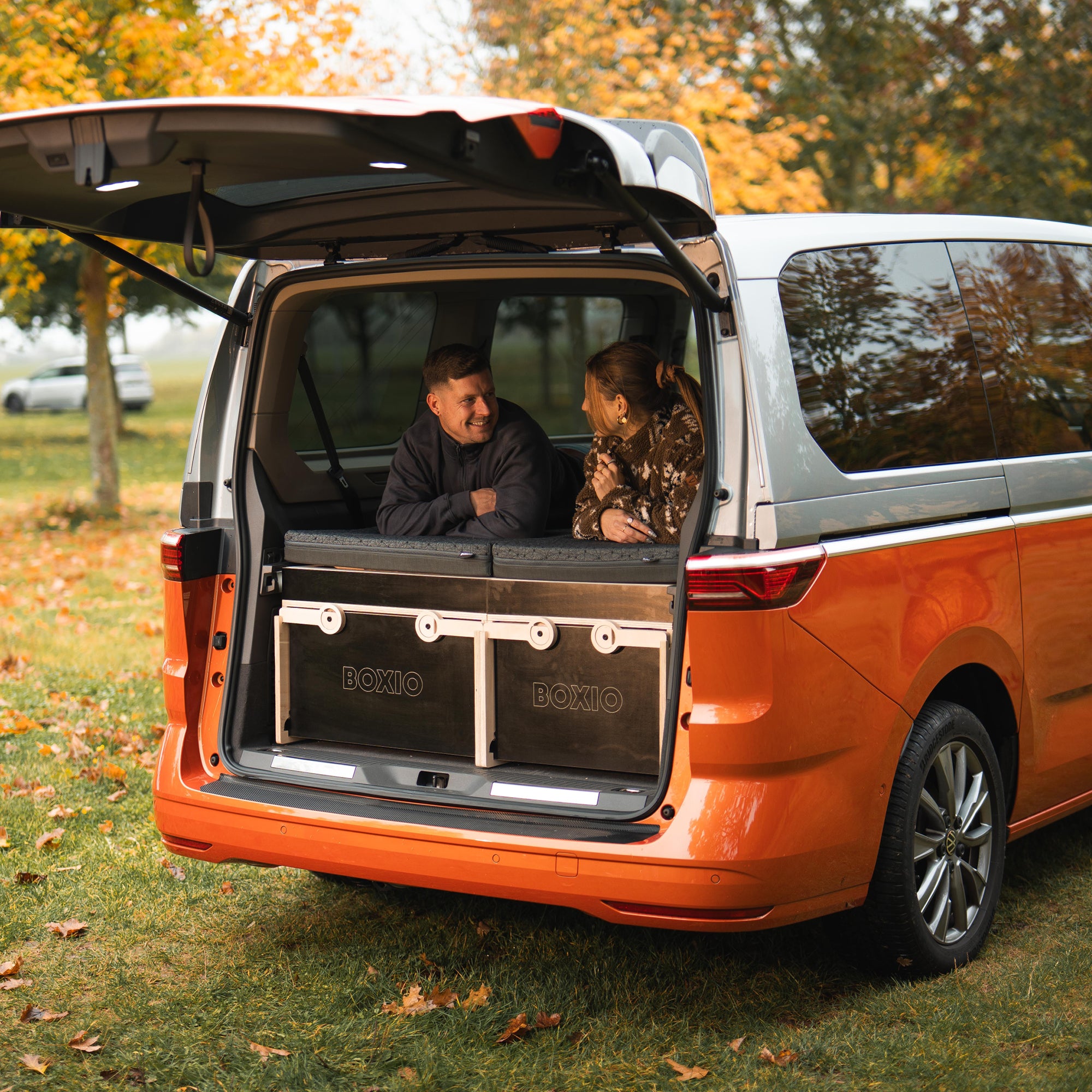 Zwei Personen sitzen in der geöffneten Heckklappe eines orangefarbenen Vans und unterhalten sich, umgeben von herbstlichen Bäumen.