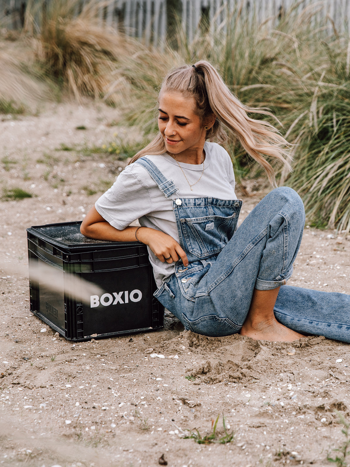 Junge Frau in blauer Jeans und T-Shirt sitzt auf dem Sand neben einer schwarzen BOXIO-Transportkiste.