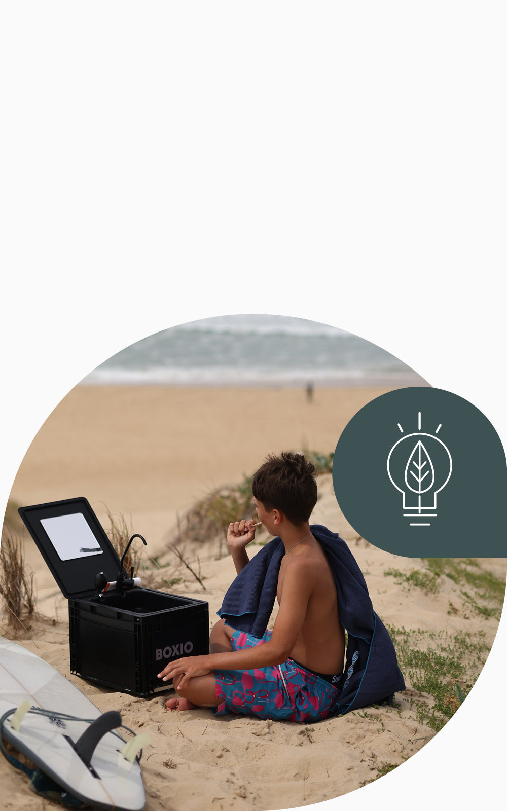 Junge im Sand sitzt neben einer schwarzen Box mit einem Spiegel, umgeben von Surfbrettern und Strand.