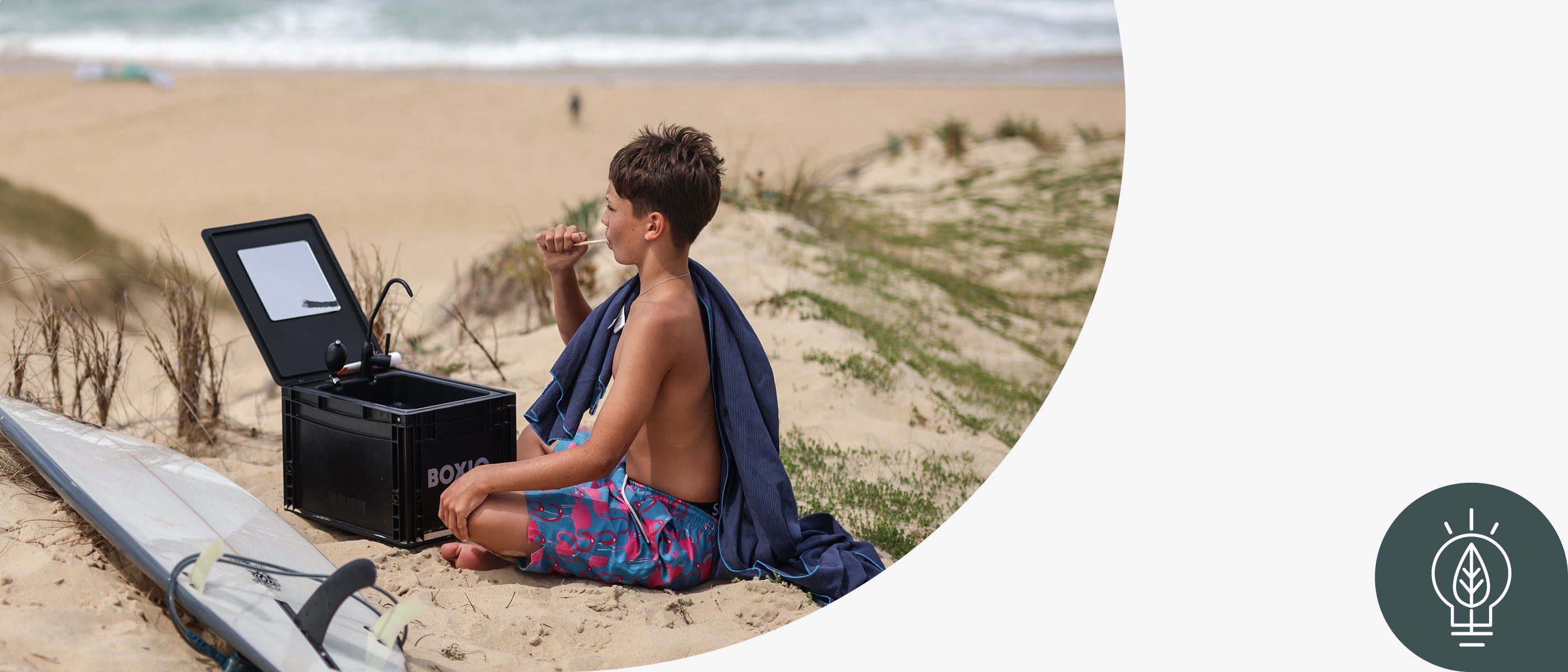 Junge sitzt am Strand, genießt eine Snacks, neben ihm ein Surfbrett und eine offene Box mit Spiegel.
