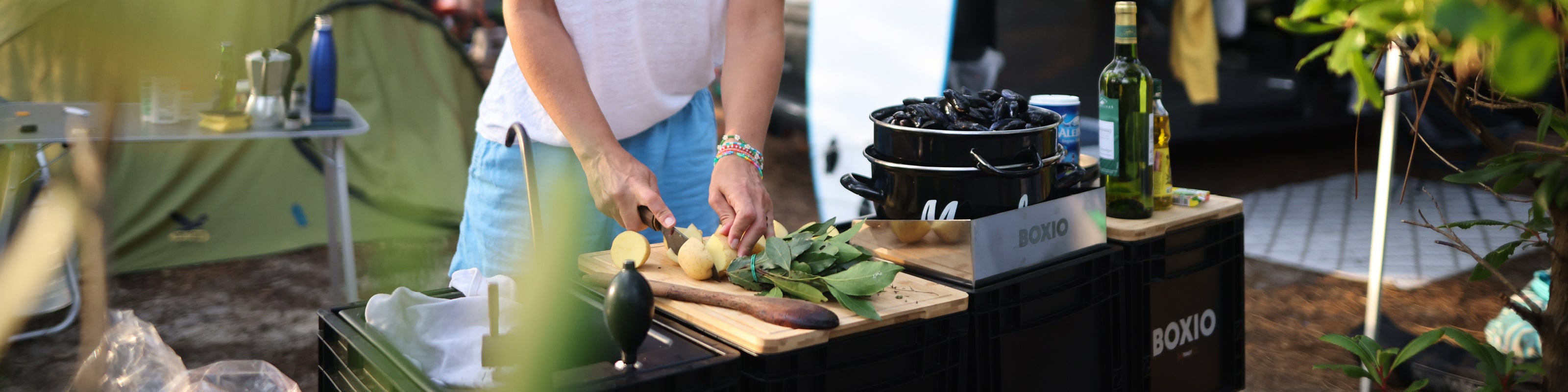 Person schneidet Gemüse, während im Hintergrund Campingausstattung und Kochutensilien sichtbar sind. Ideal für Outdoor-Kochen.