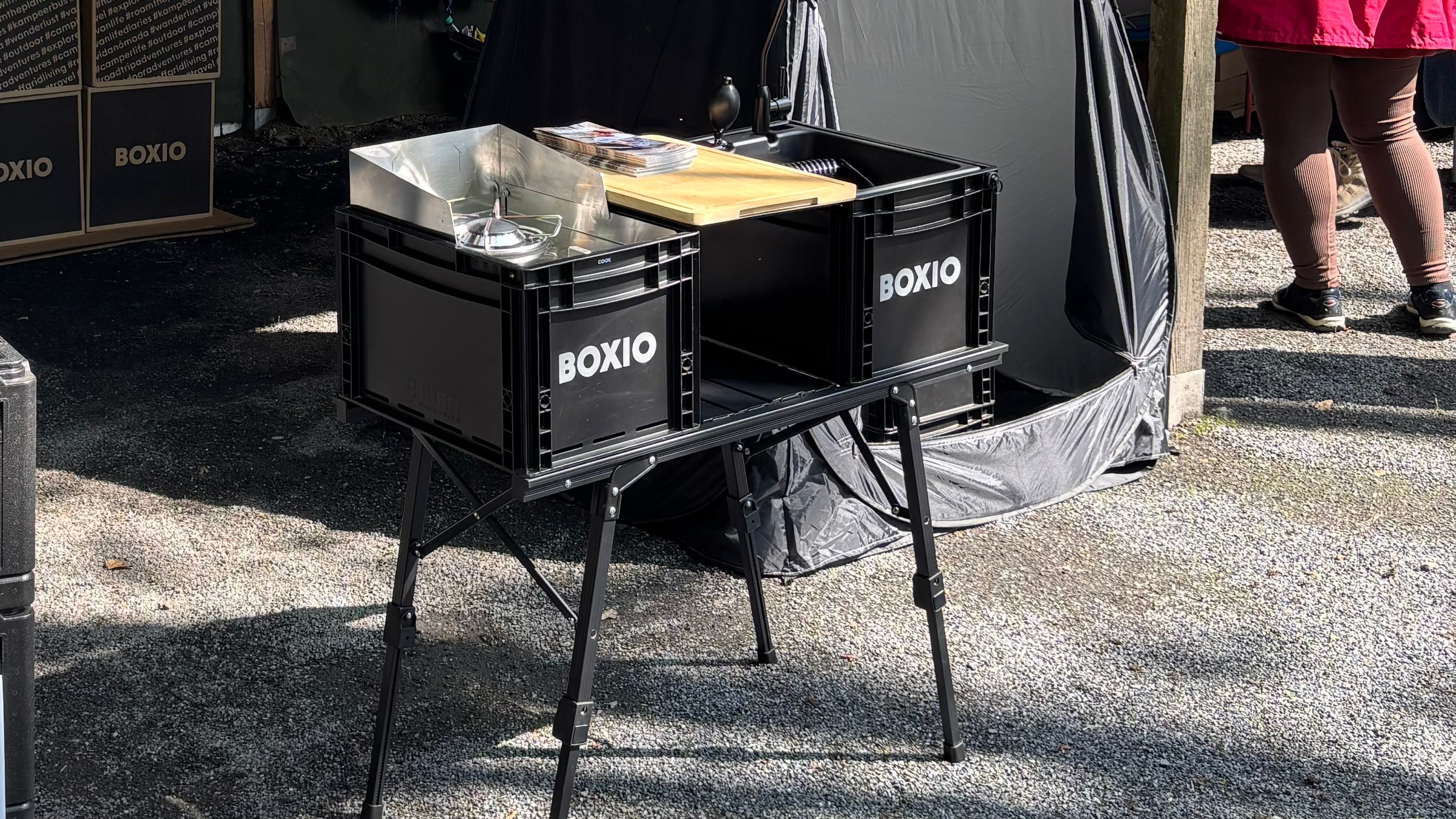 Funktionale Campingküche auf einem Stativ mit zwei schwarzen Boxen und nötigem Kochzubehör, umgeben von einer Outdoor-Umgebung.