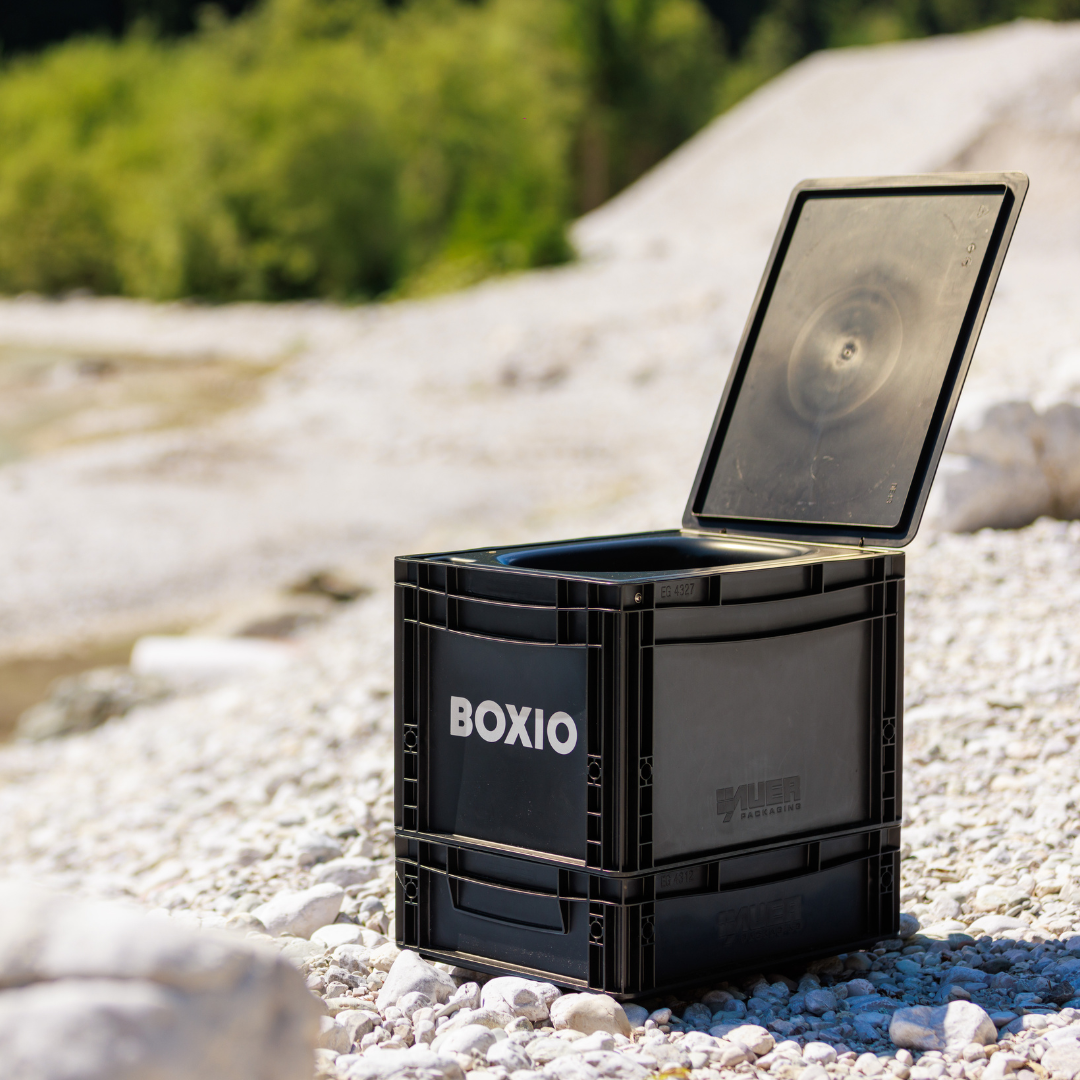 Schwarze, offene Boxio-Transportbox auf Kiesstrand, im Hintergrund grüne Bäume. Ideal für vielseitige Anwendungen.