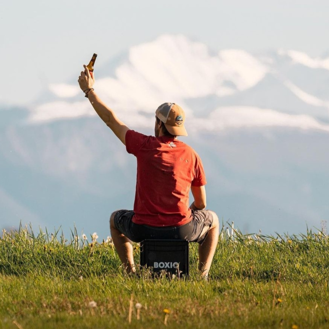 Mann sitzt auf einer Box im Gras und hebt eine Flasche, im Hintergrund schneebedeckte Berge.