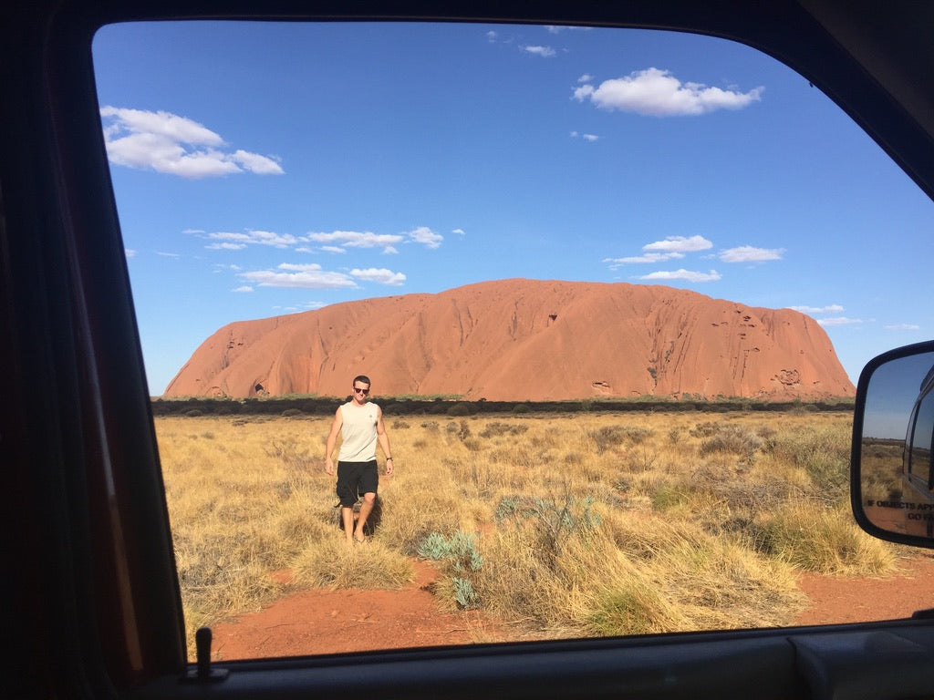 Mann steht im Outback vor dem Uluru, umgeben von bewachsenen Flächen und blauem Himmel.