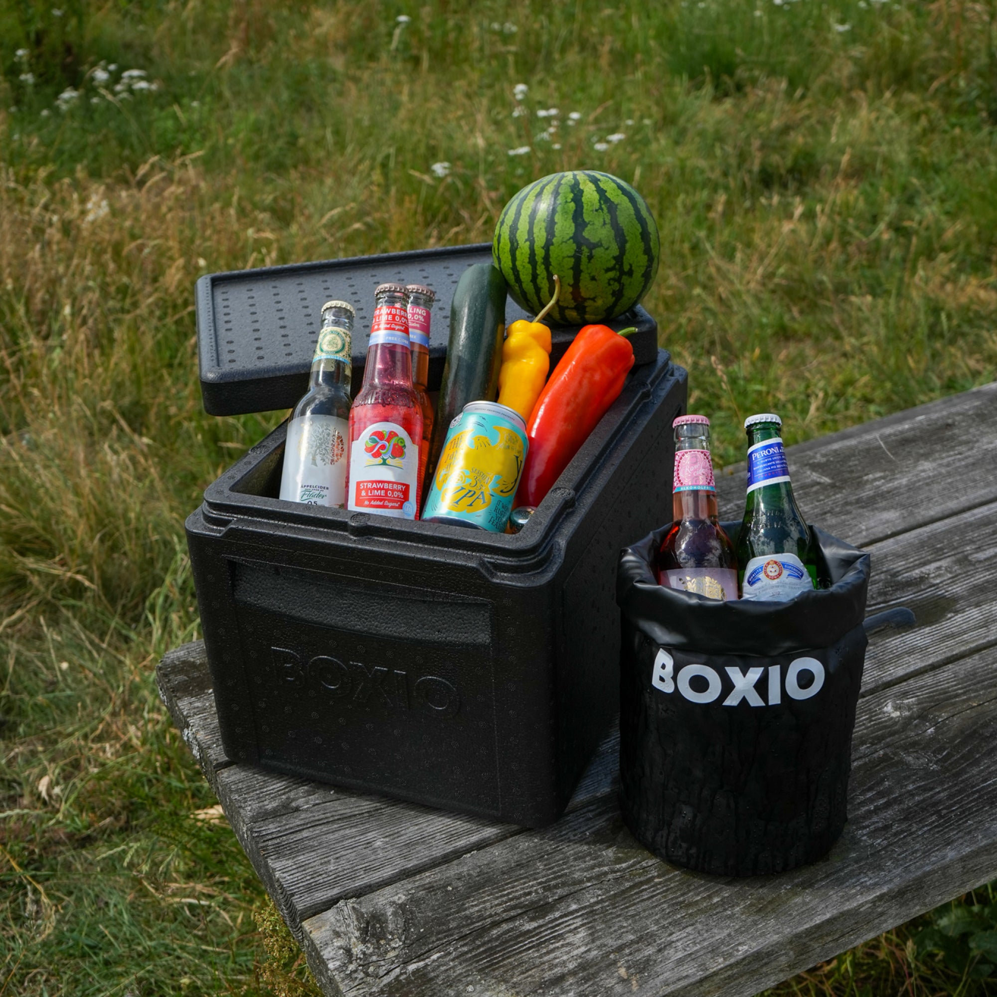 Kühlbox mit Getränken und Gemüse, darunter eine Melone, auf einer Holzbank in einer Wiese.