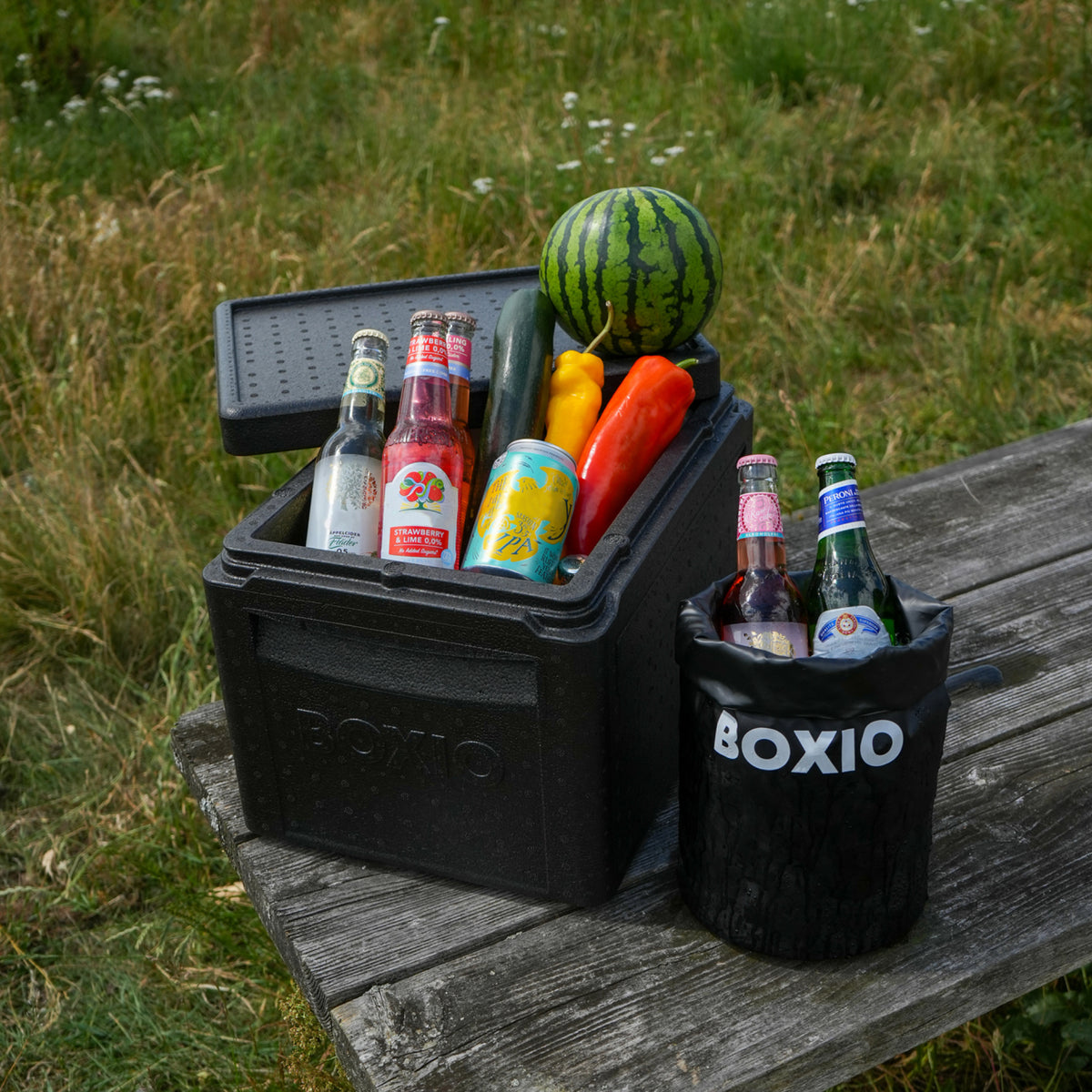 Kühlbox mit Getränken, Gemüse und einer Wassermelone, auf einem Holztisch in einer grünen Wiese.