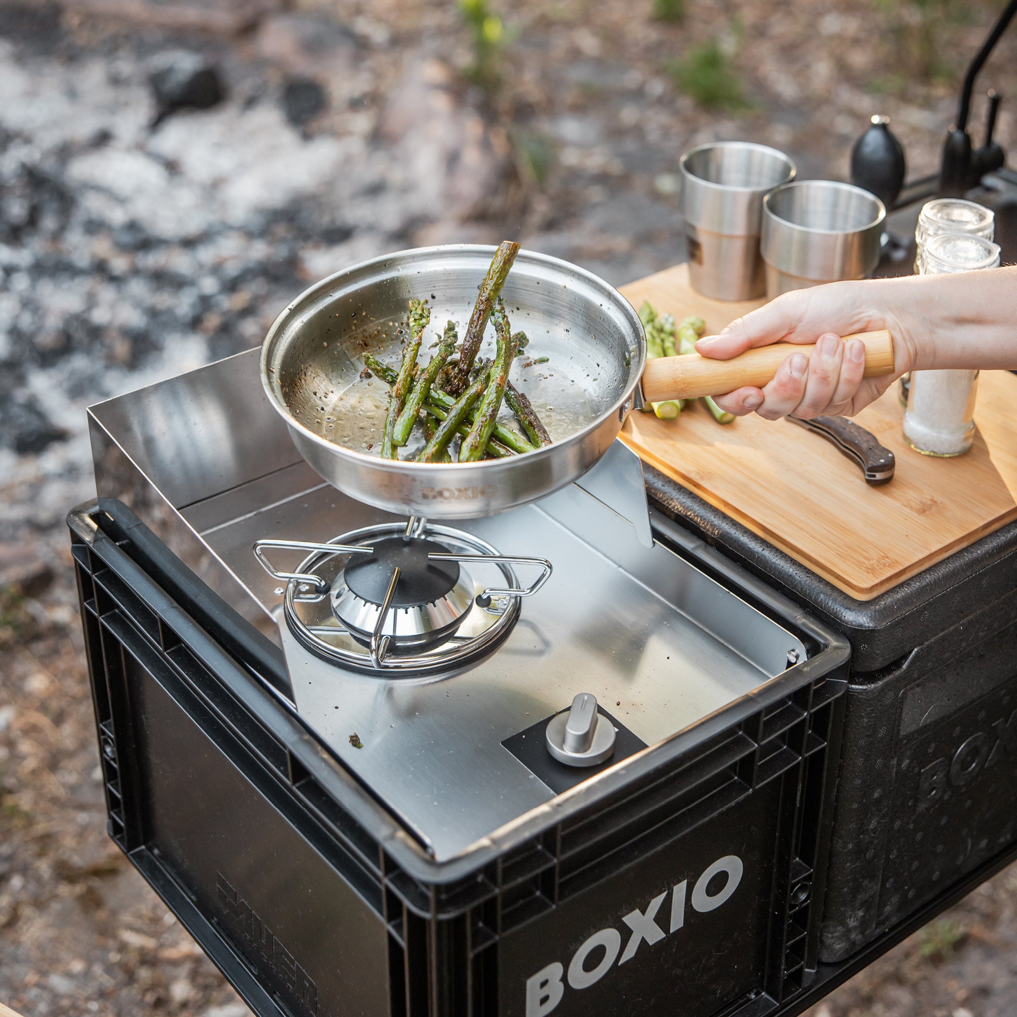 Outdoor, drei Euroboxen nebeneinander, Fokus auf dem mobilen Gaskocher darüber wird eine Edelstahlpfanne mit Holzgriff gehalten in dem Spargel gebraten wird 