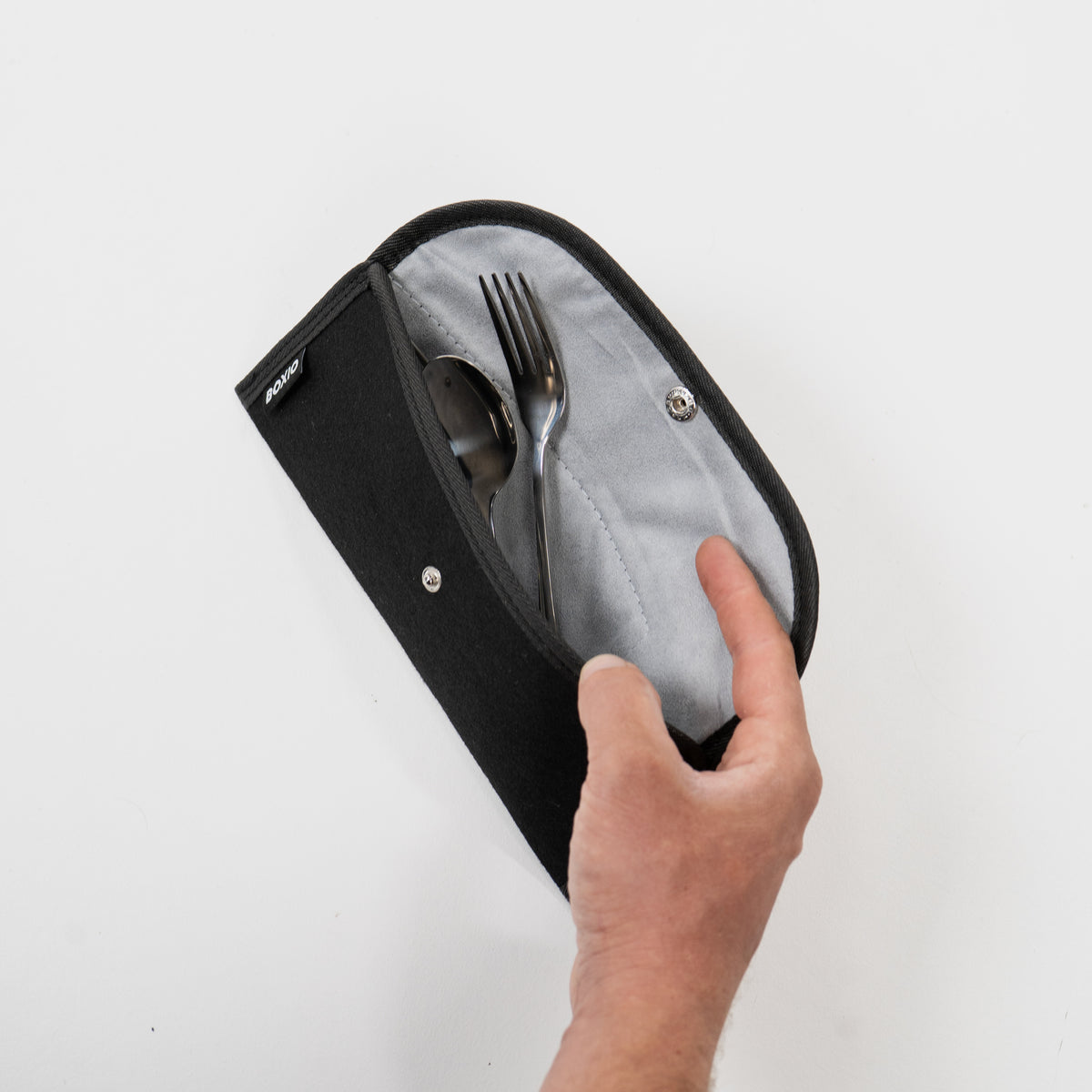 Schwarze Bestecktasche mit Besteck (Löffel und Gabel) und heller Innenausstattung, Hand öffnet die Tasche.