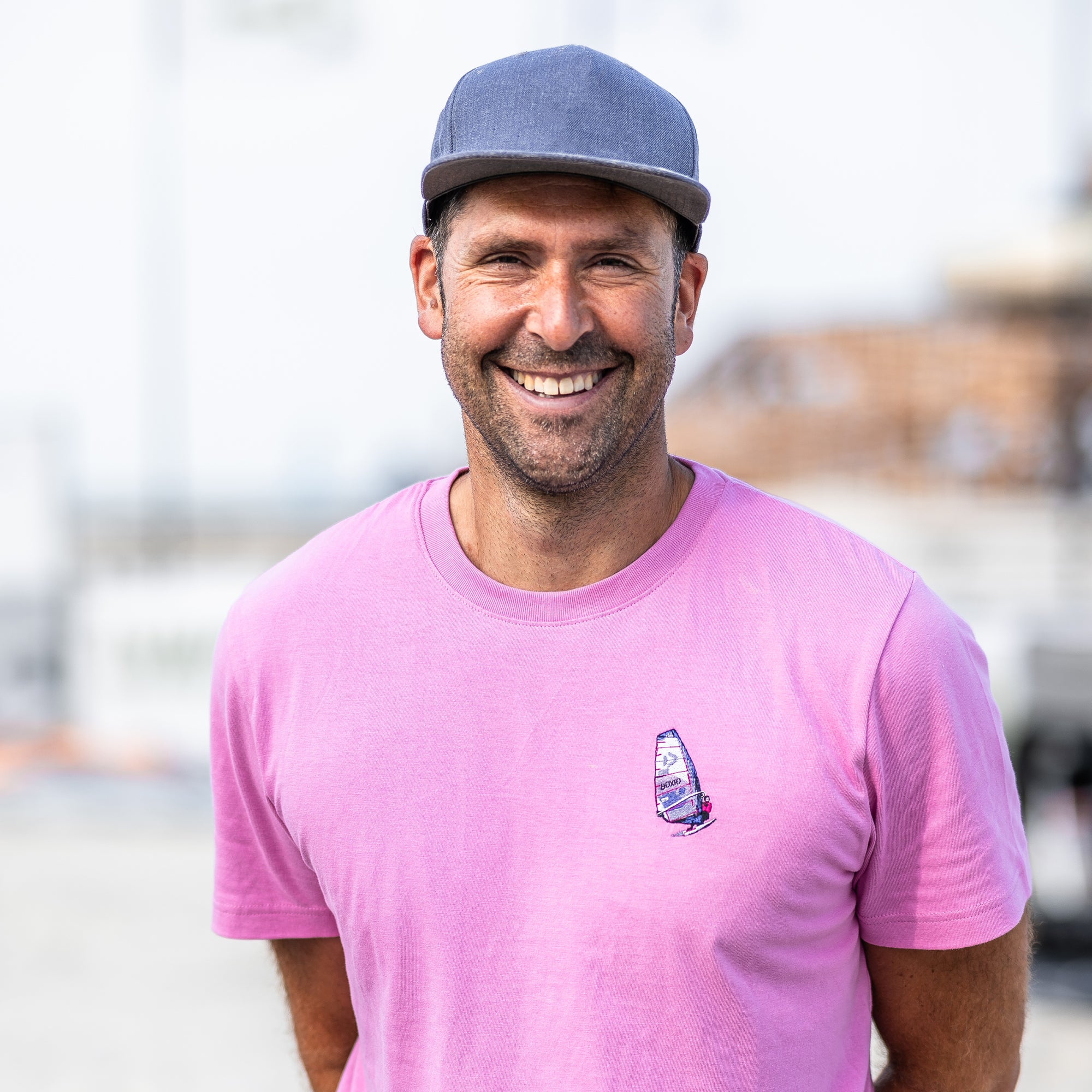 Lachender Mann in pinkem T-Shirt mit Segelboot-Motiv und blauer Kappe, vor unscharfem Hintergrund.
