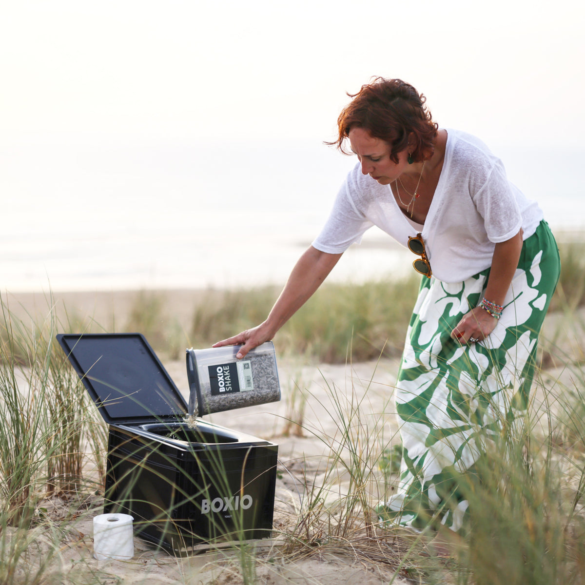 Frau im grünen Rock und weißem Oberteil füllt einen Behälter in eine Kühlbox am Strand.