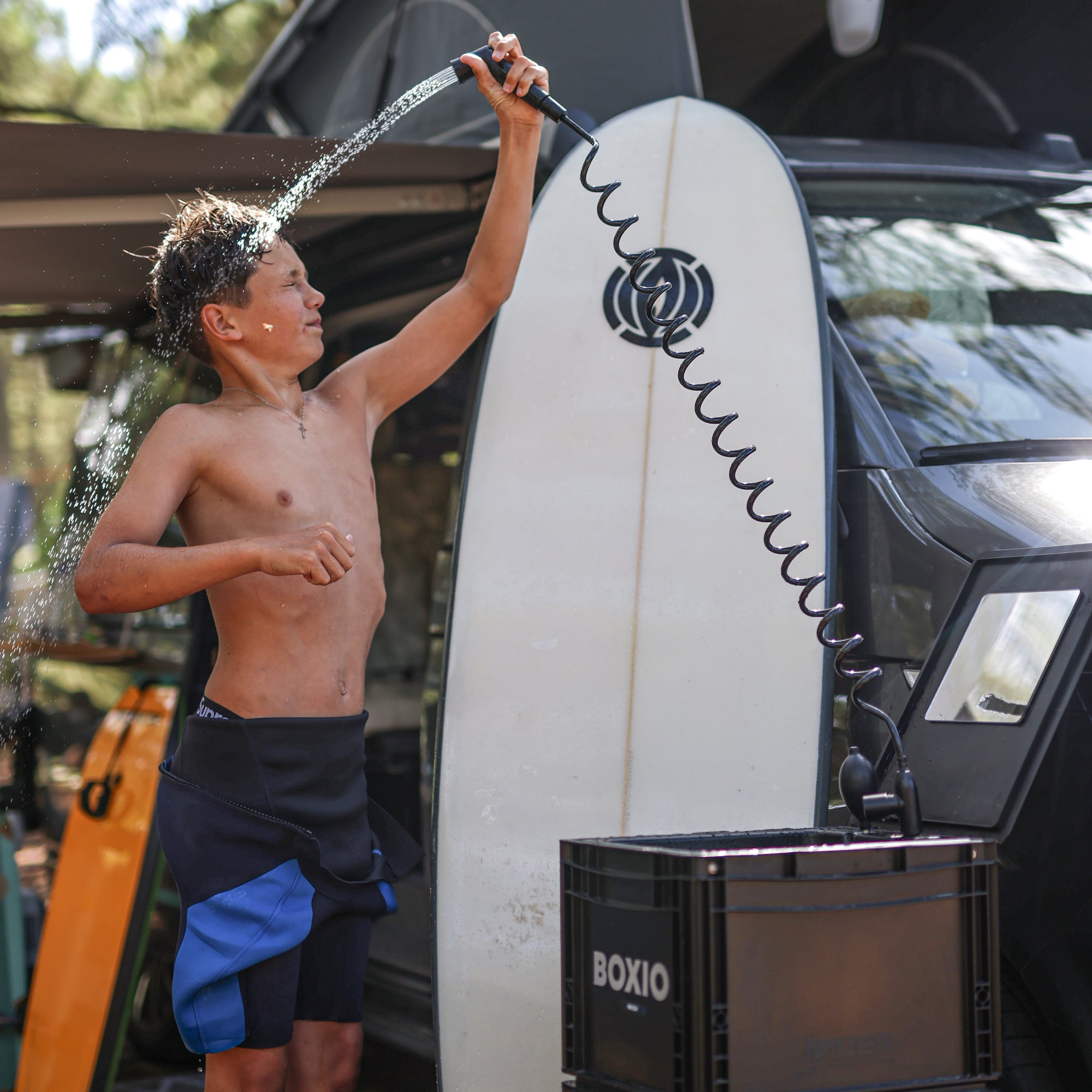 Junge spritzt sich mit einem Schlauch Wasser über den Körper, im Hintergrund ein Surfbrett und ein Wohnmobil.