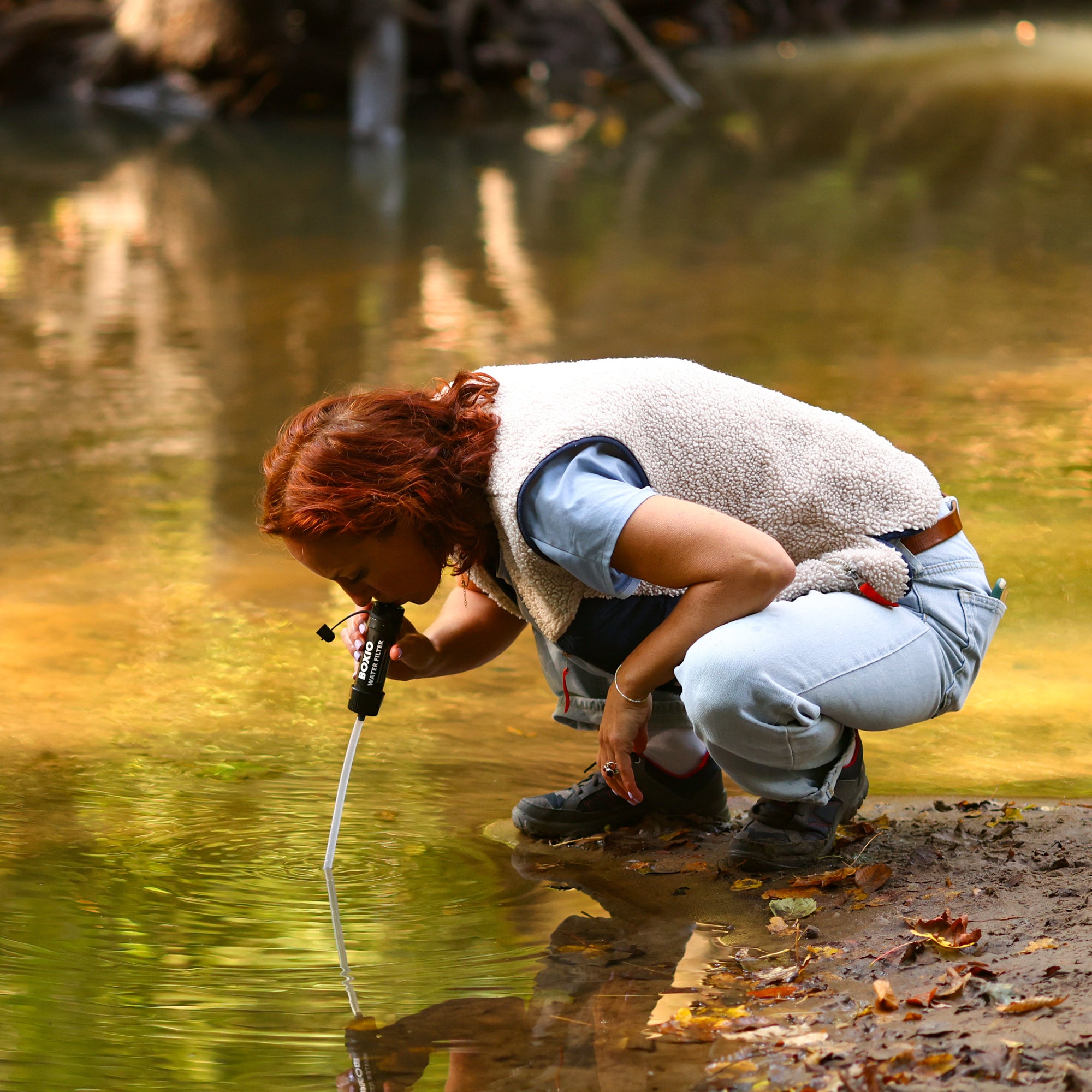 Frau in einem Gewässer untersucht mit einem Messinstrument die Wasserqualität, umgeben von natürlicher Vegetation.