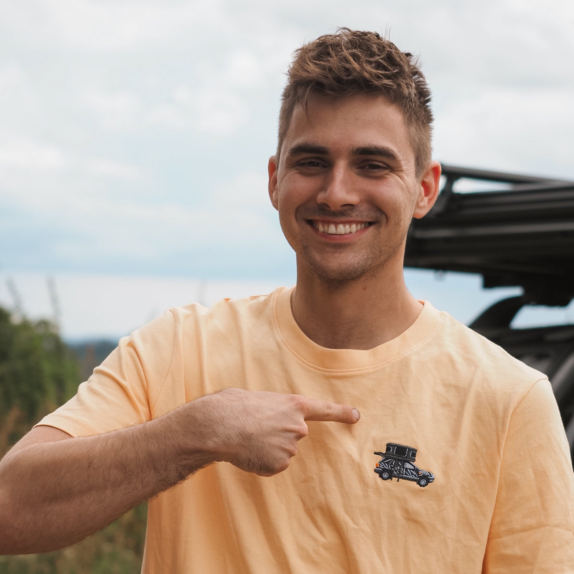 Junger Mann in gelbem T-Shirt zeigt lächelnd auf ein Auto-Motiv auf der Brust, vor einer Landschaft mit Bäumen.