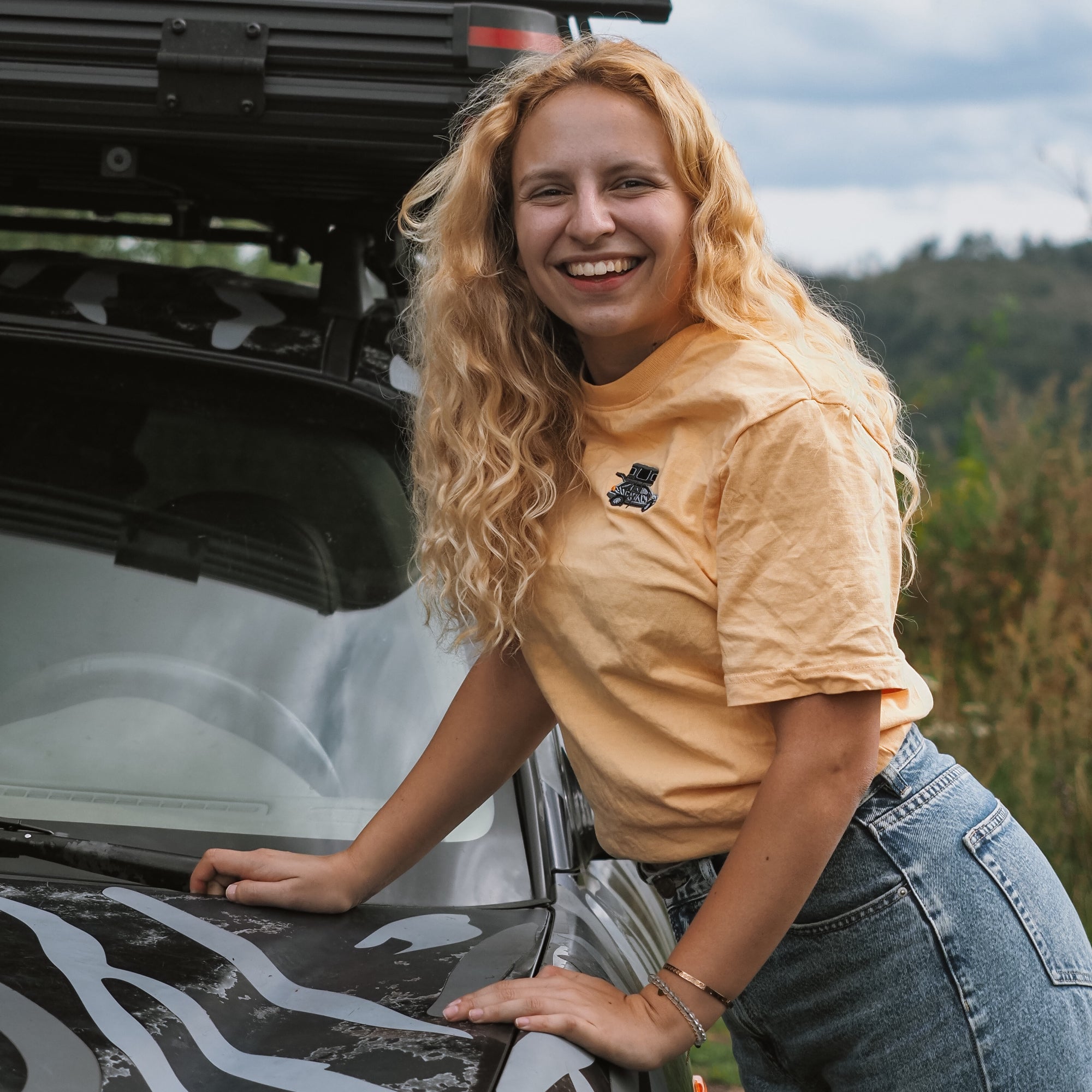 Lächelnde junge Frau mit lockigem, blondem Haar lehnt auf einem Auto und trägt ein gelbes T-Shirt und Jeans.