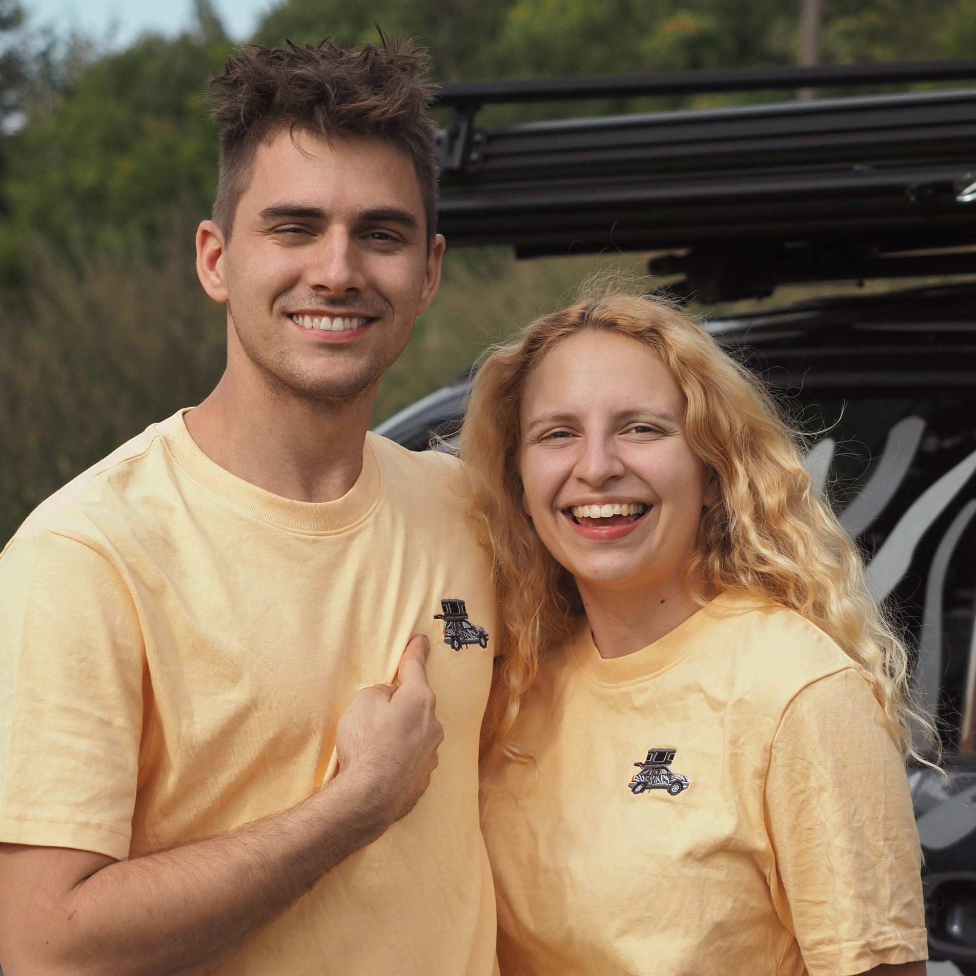 Lächelndes Paar in gelben T-Shirts mit Aufdruck, umgeben von Natur und einem Fahrzeug im Hintergrund.