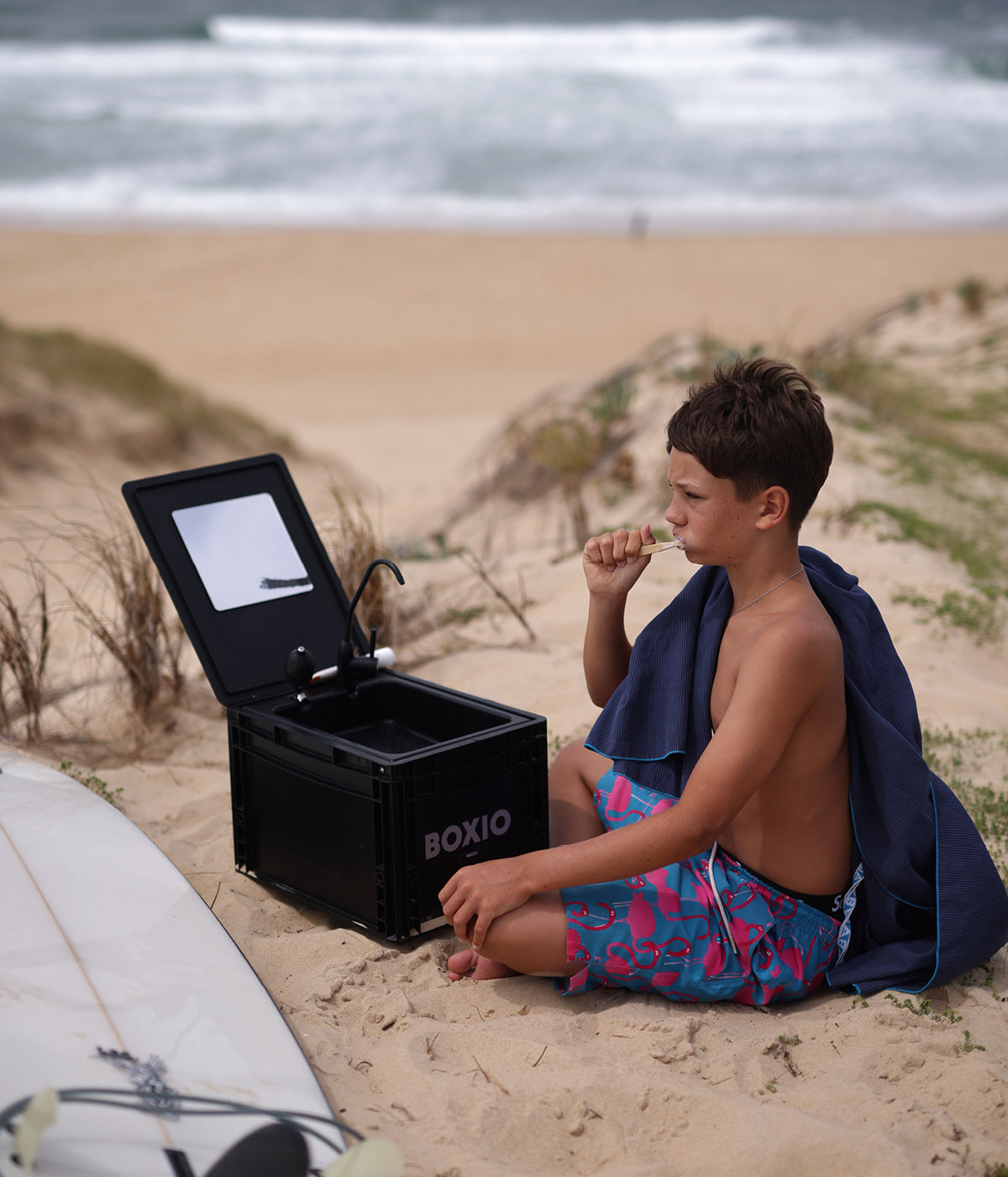Junge im Sand am Strand sitzt neben schwarzer BOXIO-Waschkiste und genießt einen Snack, mit Surferbrett im Vordergrund.