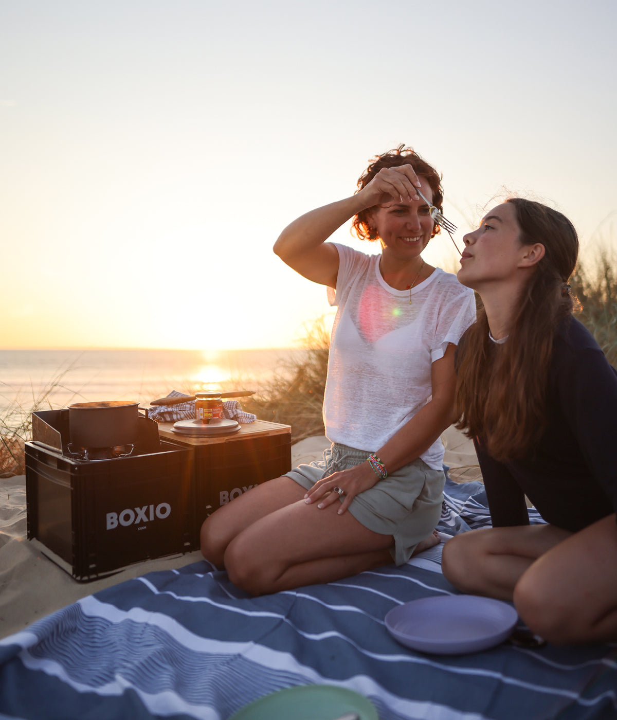 Zwei Frauen genießen ein Abendessen am Strand bei Sonnenuntergang, umgeben von Picknickutensilien und einer Decke.
