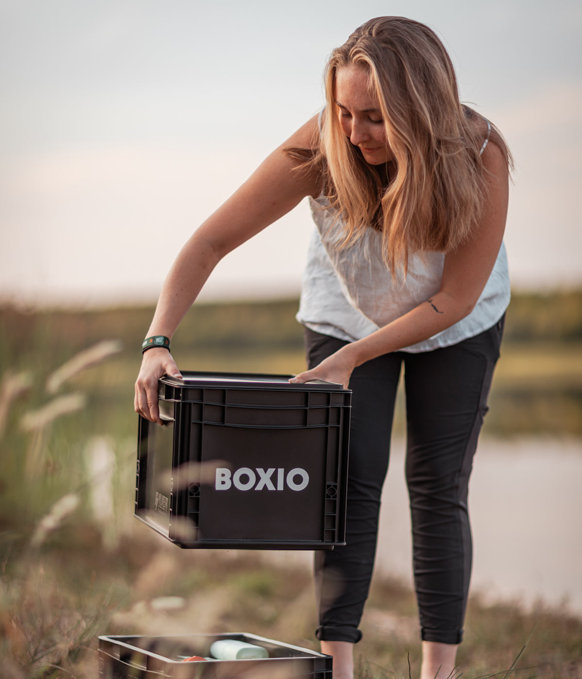 Frau hebt eine schwarze Box mit der Aufschrift "BOXIO" am Ufer eines Gewässers.