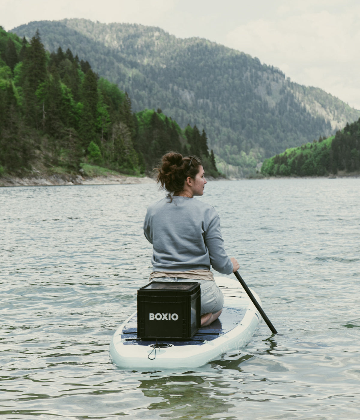 Frau paddelt auf einem Stand-Up-Paddle-Board in einem ruhigen See, umgeben von grünen Bergen und Bäumen.