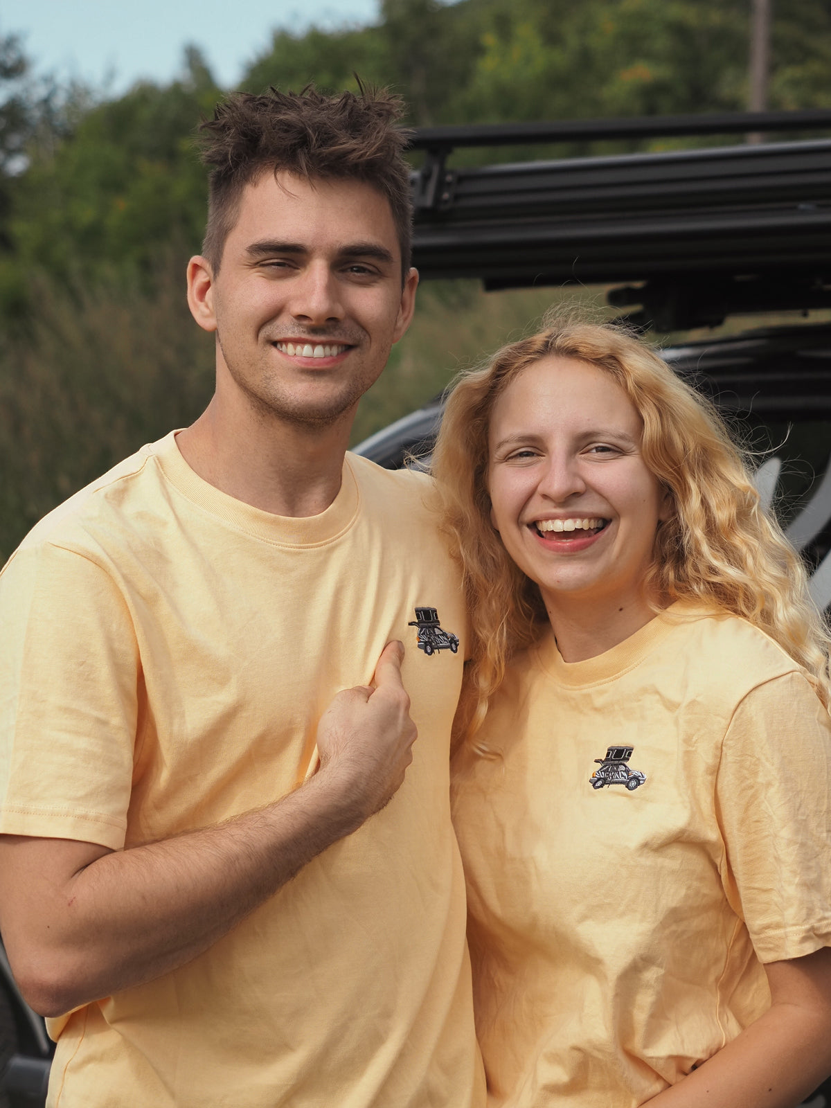 Zwei Personen mit blonden Haaren tragen gelbe T-Shirts mit einem Auto-Logo, stehen lächelnd vor einem Auto im Freien.