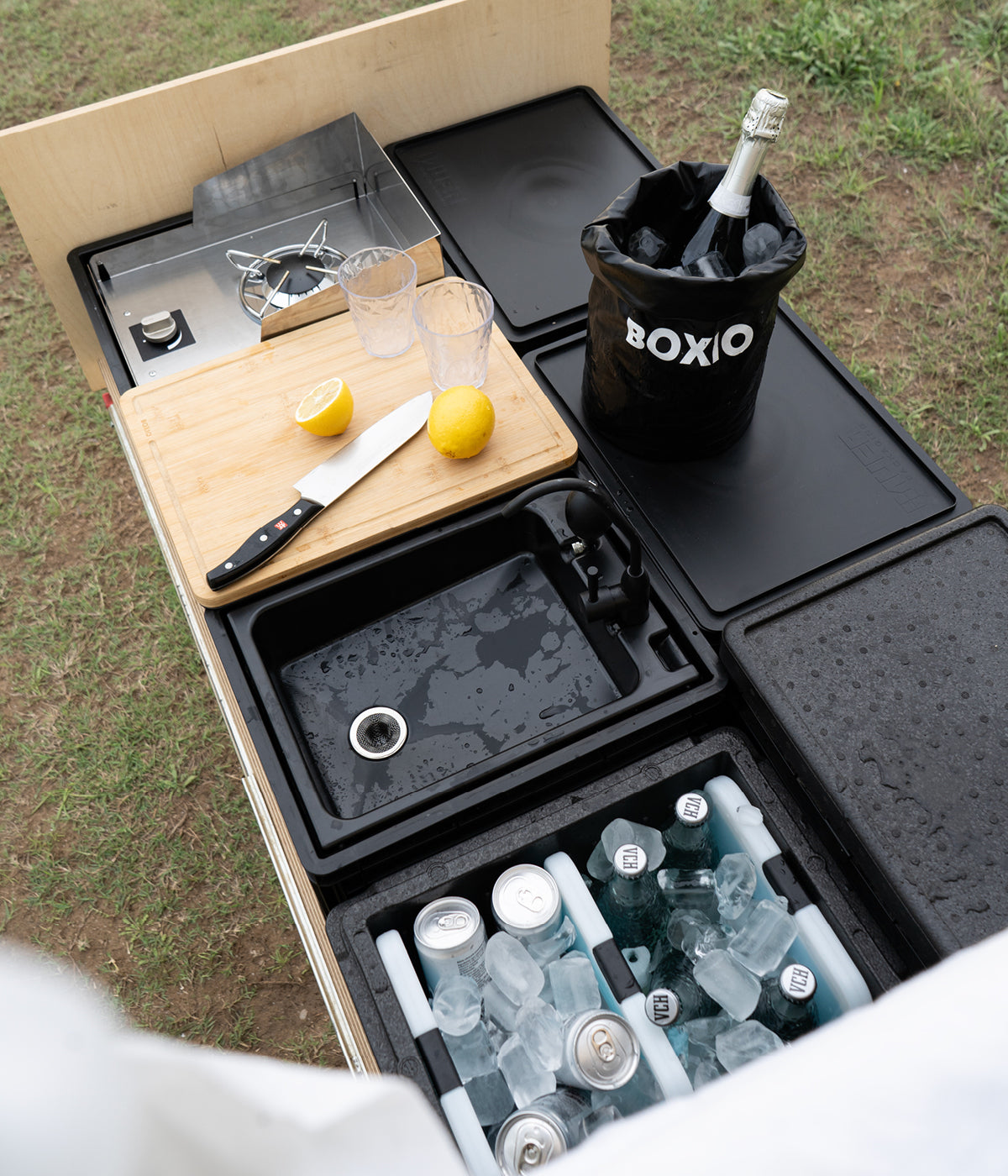 Küchenbereich im Freien mit Kocher, Schneidebrett, Glasbehältern, Zitronen und gekühlten Getränken in einer Kühlbox.