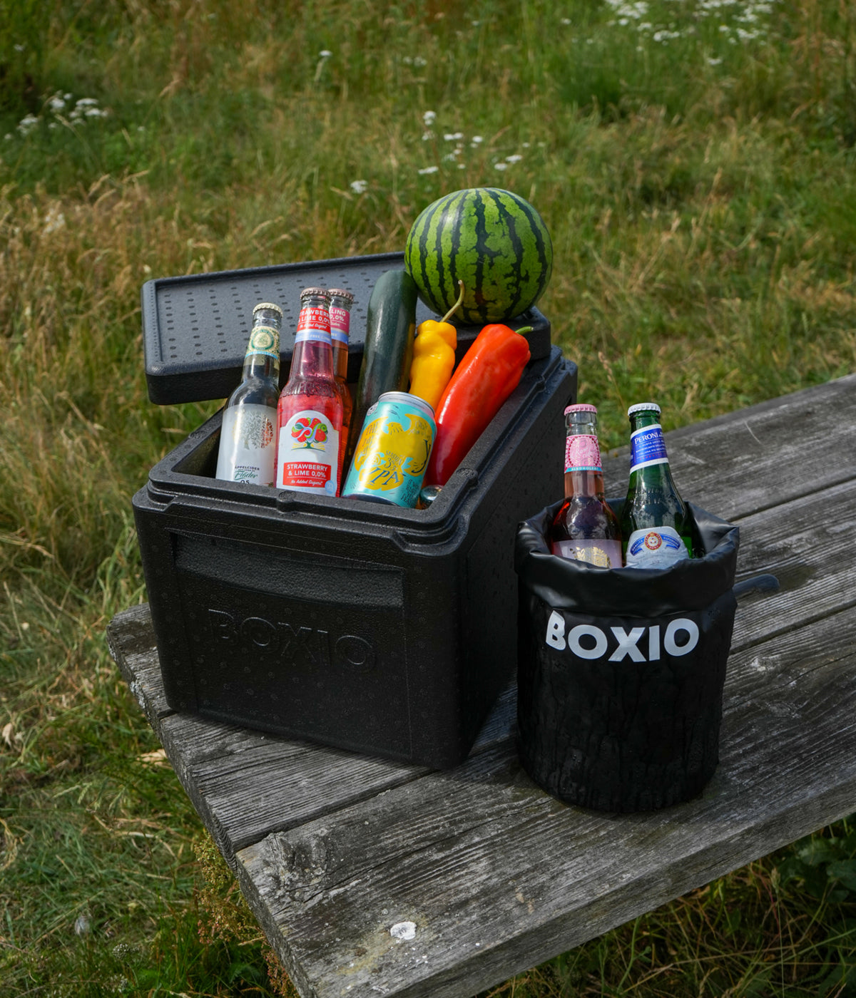 Kühlbox mit Getränken, Gemüse und einer Wassermelone auf einer Holztischplatte im Freien.
