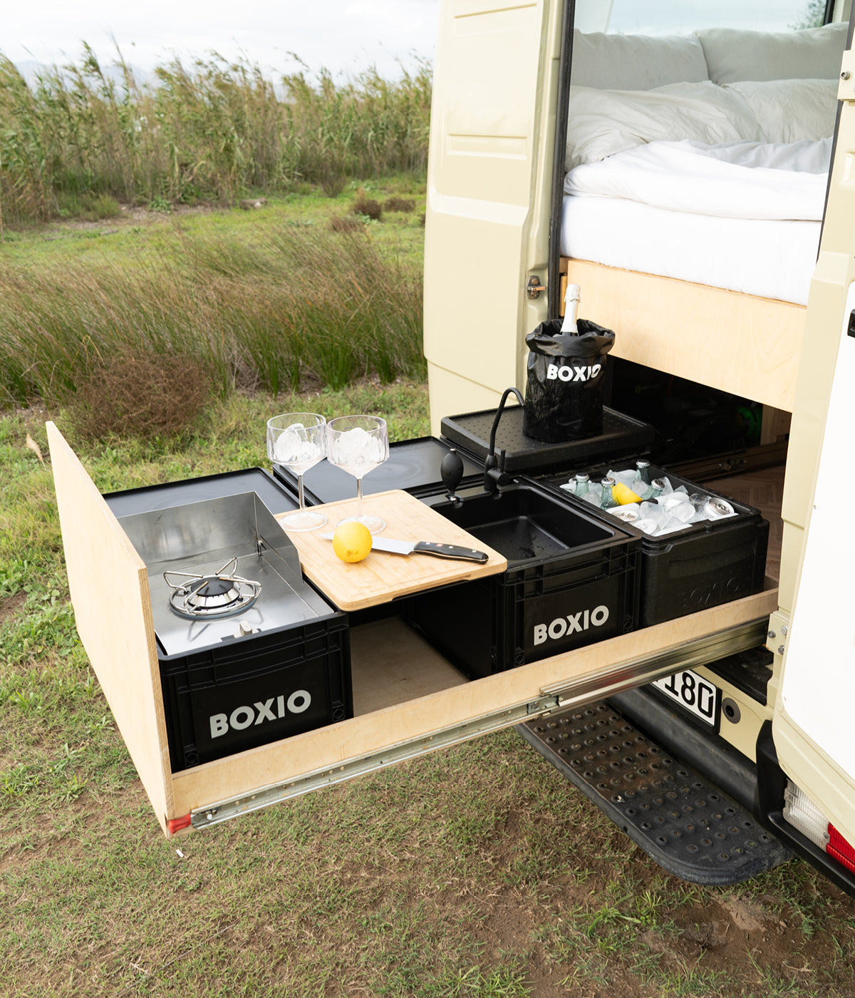 Mobile Küchenzeile eines Campervans mit Kocher, Gläsern, Schneidebrett und frischen Zutaten im Freien.