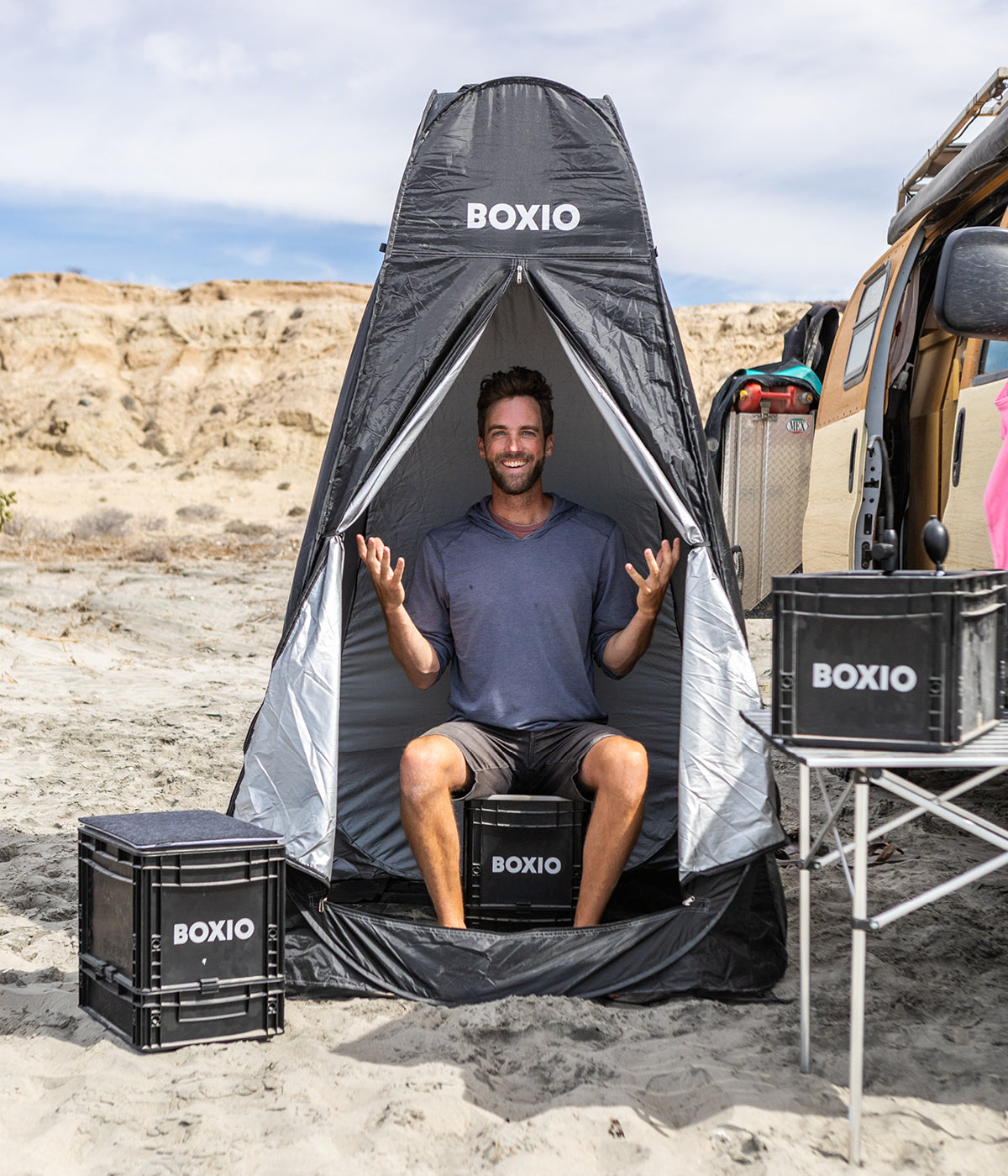 Ein Mann sitzt entspannt in einem schwarzen Portabler Raum auf einem Boxio-Kasten, umgeben von Sand und einem Van.