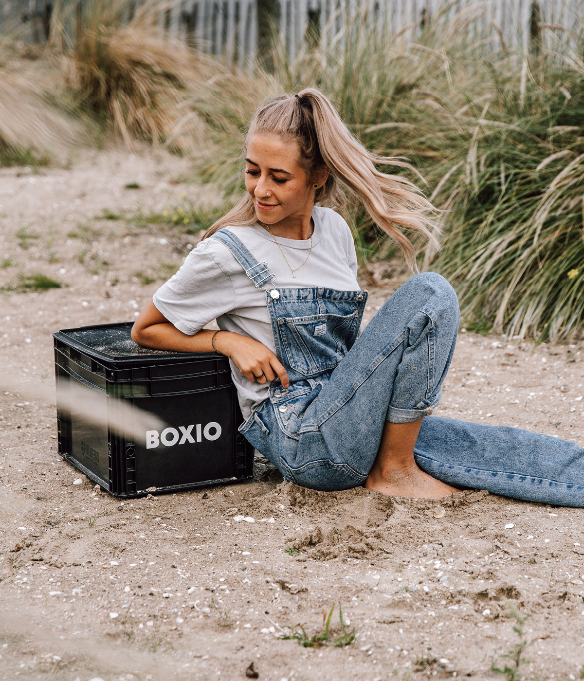 Junge Frau in Latzhose sitzt auf dem Boden, neben einer schwarzen Box mit der Aufschrift "BOXIO", umgeben von Sand und Gras.